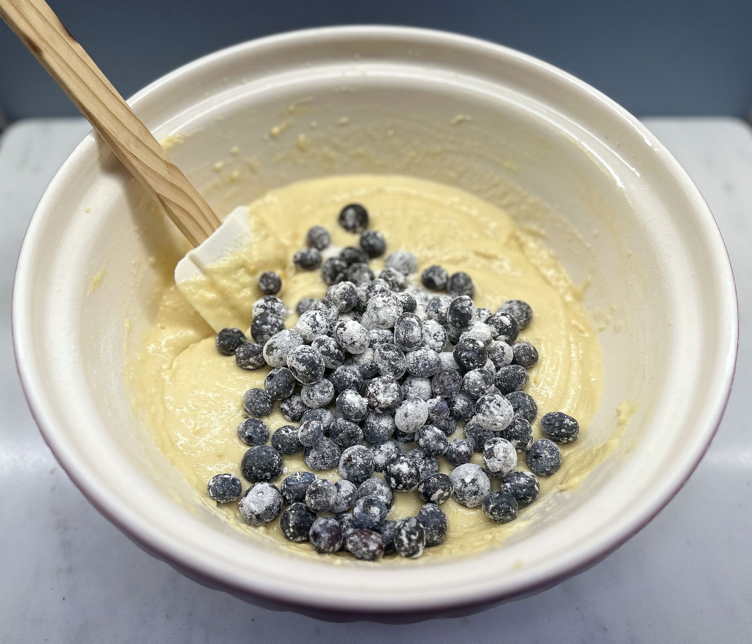 A mixing bowl filled with a smooth, yellow cake batter. A pile of blueberries, lightly dusted with flour, is placed on top of the batter. A spatula rests in the bowl, suggesting that the blueberries are about to be gently folded into the batter. The flour coating on the blueberries helps prevent them from sinking to the bottom during baking, ensuring an even distribution throughout the cake.