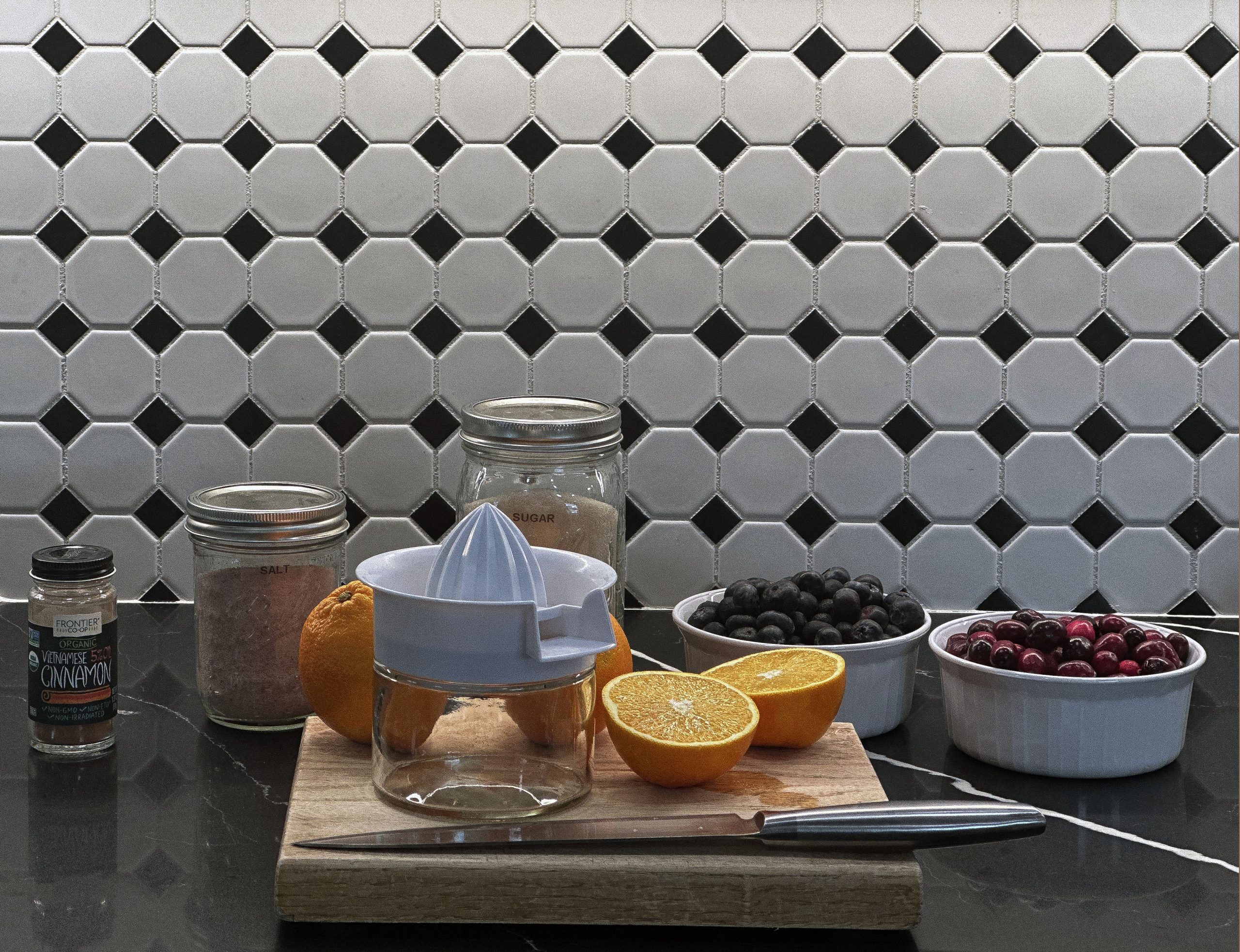 Ingredients for cranberry-blueberry sauce, including fresh blueberries, cranberries, oranges, sugar, salt, and cinnamon, arranged on a black countertop with a tiled background.