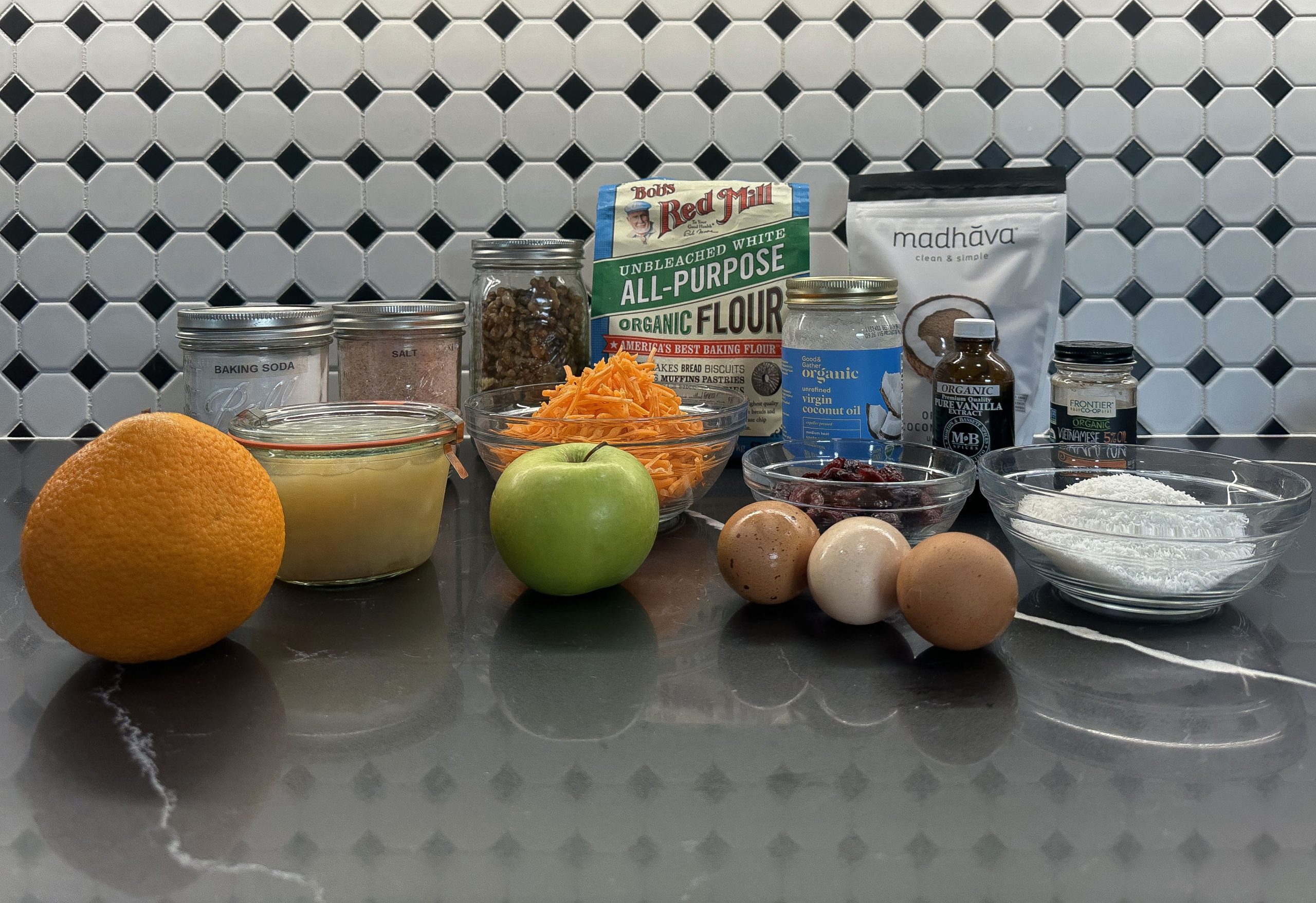 A collection of ingredients for Cranberry Morning Glory Muffins displayed on a kitchen counter. Ingredients include an orange, a green apple, shredded carrots, eggs, dried cranberries, coconut flakes, walnuts, flour, coconut oil, applesauce, cinnamon, vanilla extract, sugar, salt, and baking soda. The ingredients are neatly arranged in bowls, jars, and packaging, set against a stylish black-and-white hexagon tile backsplash. 
