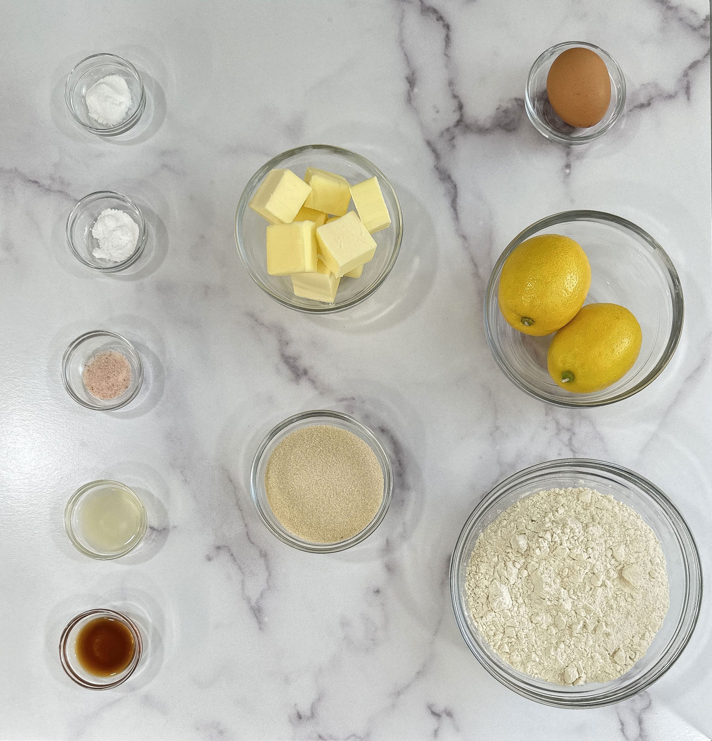 An overhead view of ingredients for lemon sugar cookies, including butter, flour, sugar, lemons, an egg, and baking essentials on a marble surface.