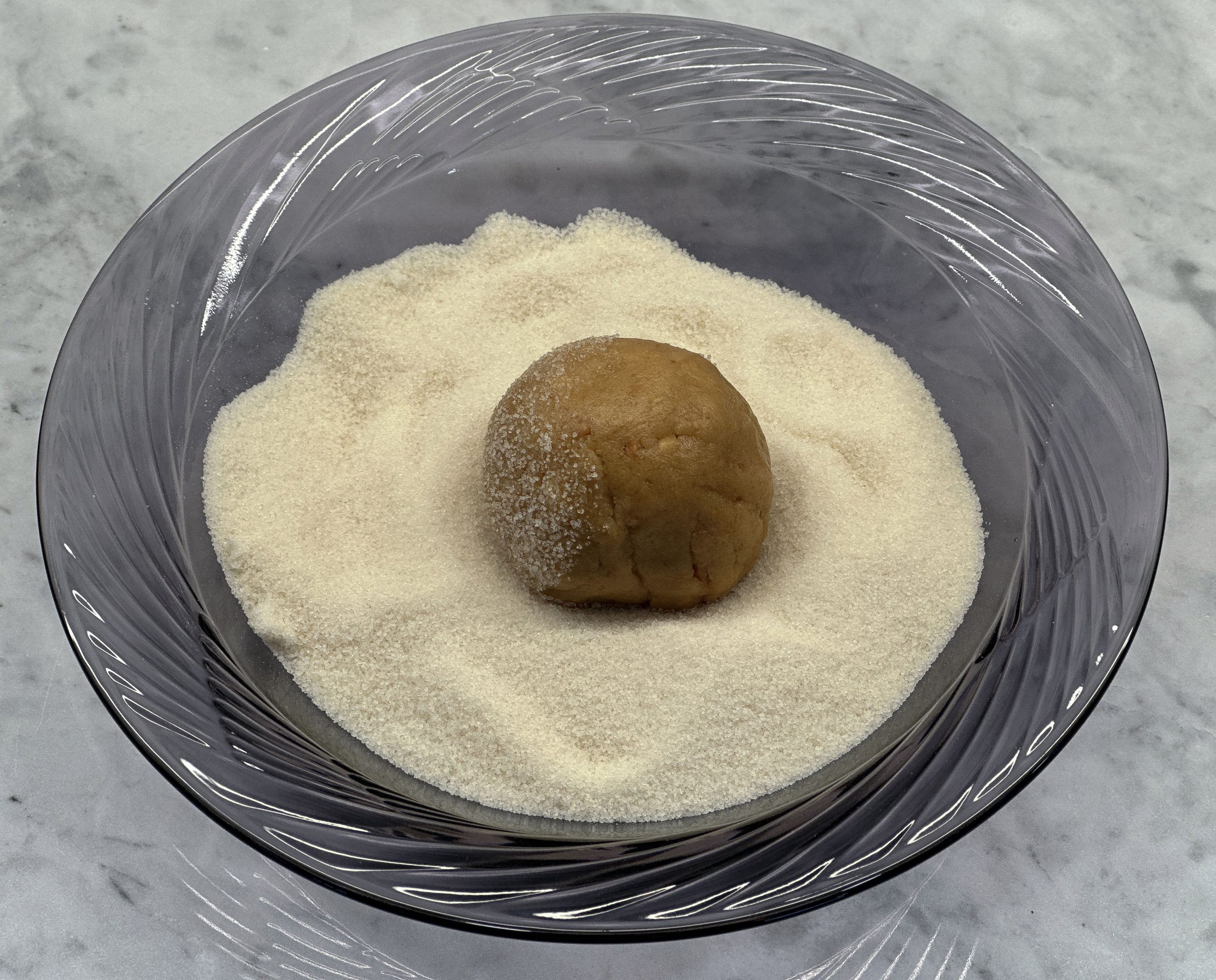 A ball of peanut butter cookie dough being rolled in a bowl of white sugar, ready for baking.