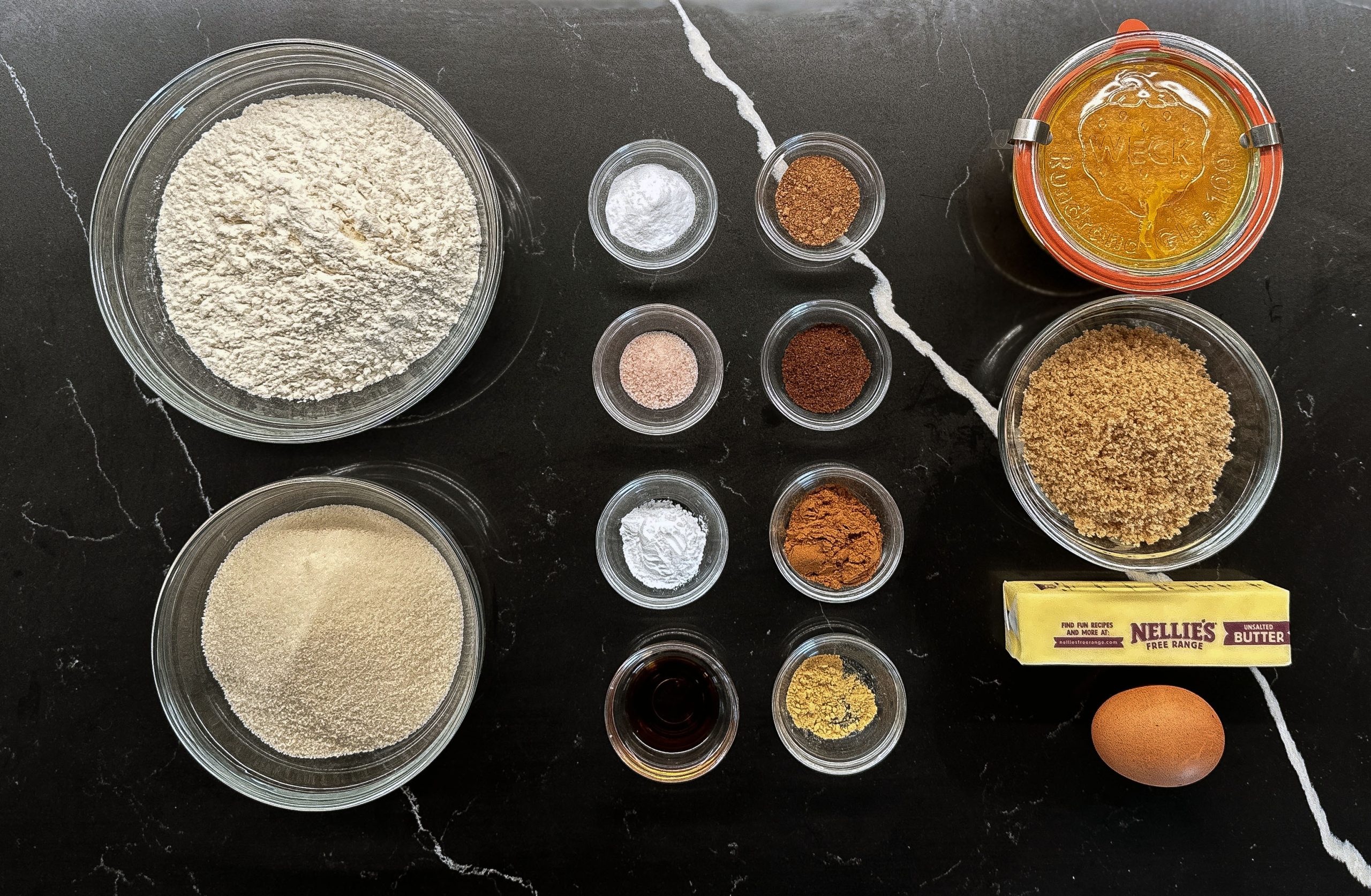 An overhead view of neatly arranged pumpkin cookie ingredients, including flour, sugars, spices, butter, and an egg, displayed on a sleek black countertop.