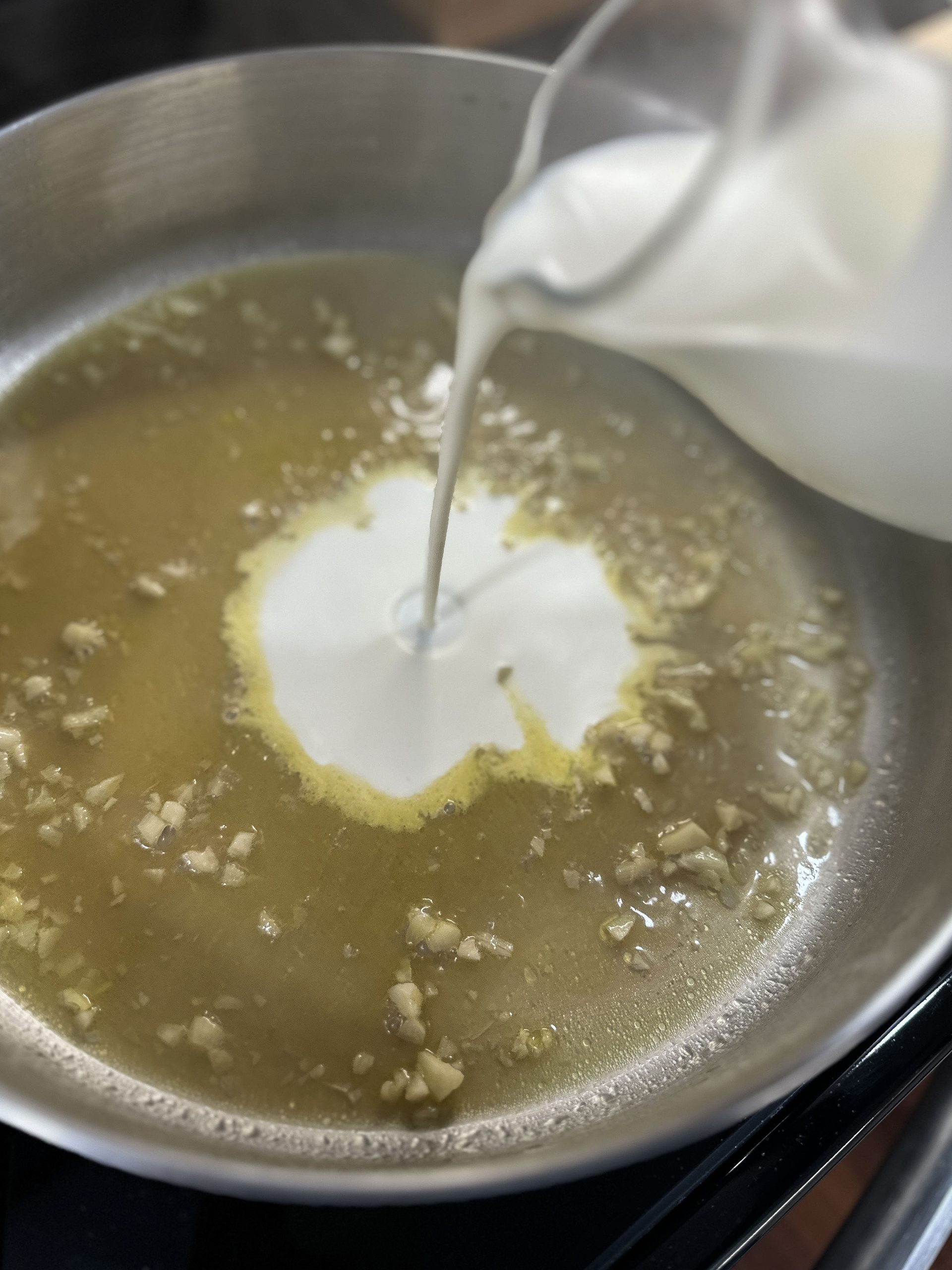 Heavy cream being poured into a garlic and broth mixture in a pan to create a creamy sauce.