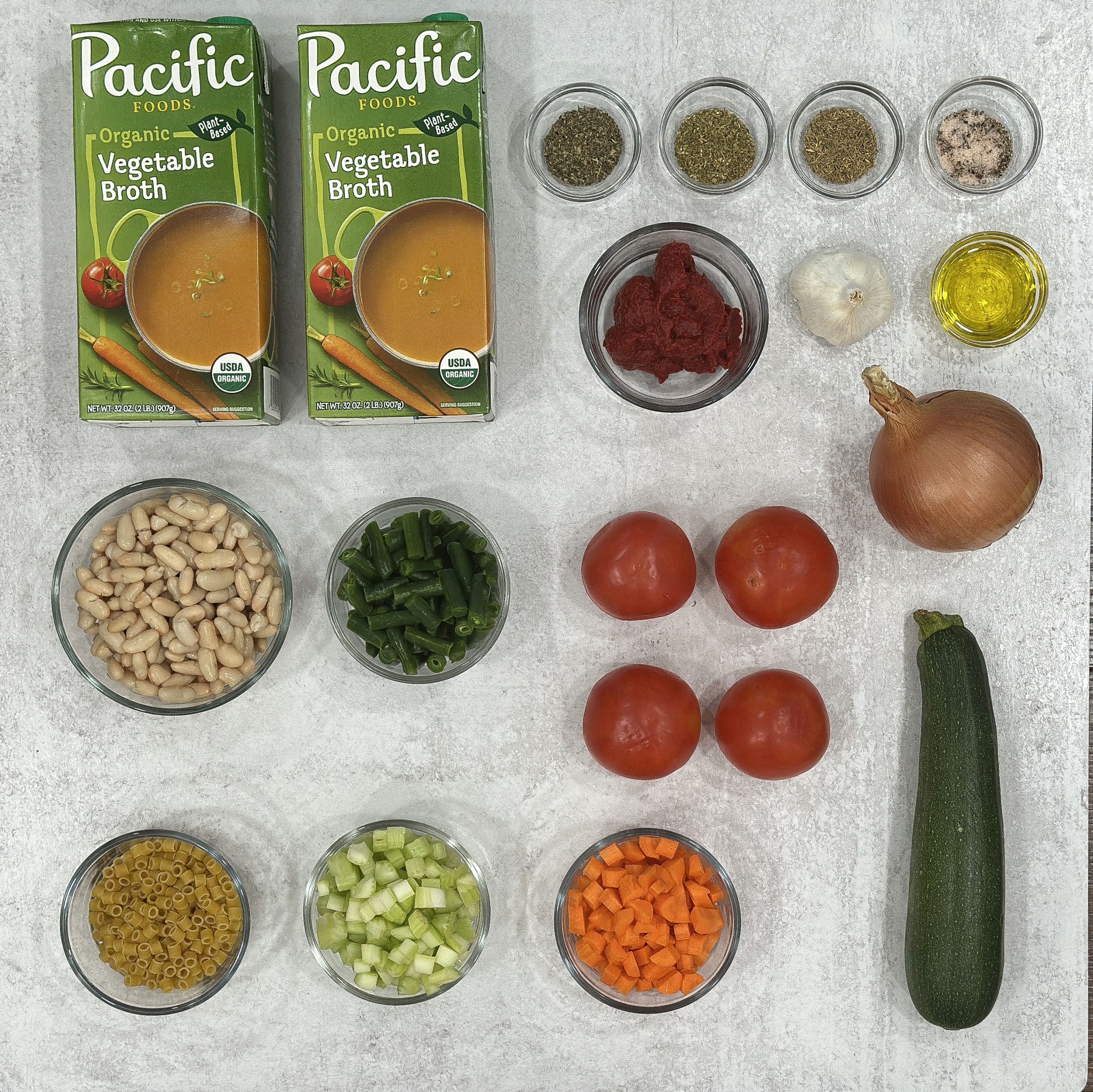 A top-down view of fresh ingredients neatly arranged for making homemade minestrone soup, including vegetables, beans, pasta, broth, and seasonings.