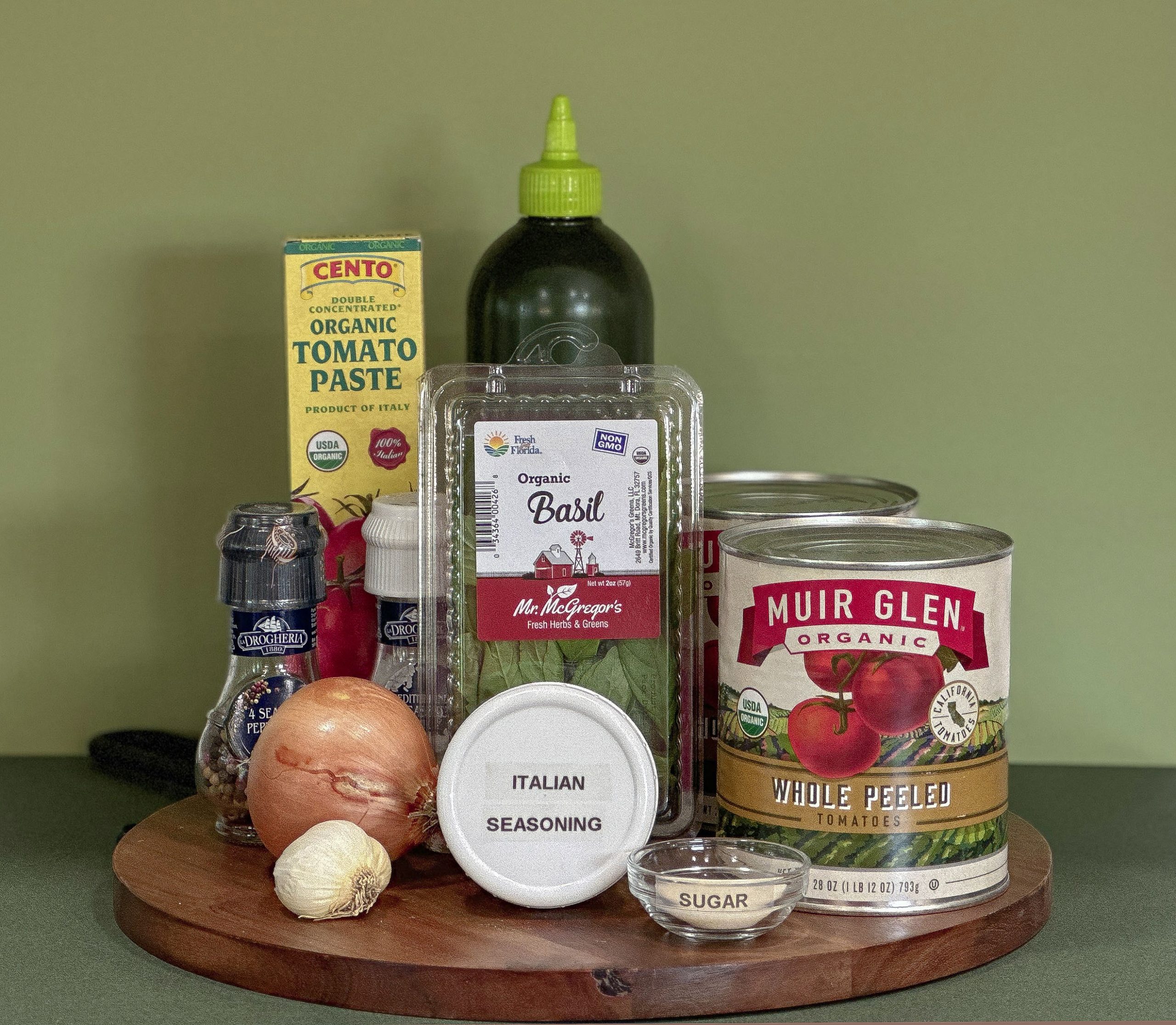 Ingredients for homemade marinara sauce arranged on a wooden board. Items include cans of Muir Glen whole peeled tomatoes, a box of Cento organic tomato paste, a container of Mr. McGregor's organic basil, an onion, a garlic bulb, a small bowl of sugar, Italian seasoning, a bottle of olive oil, and salt and pepper grinders.