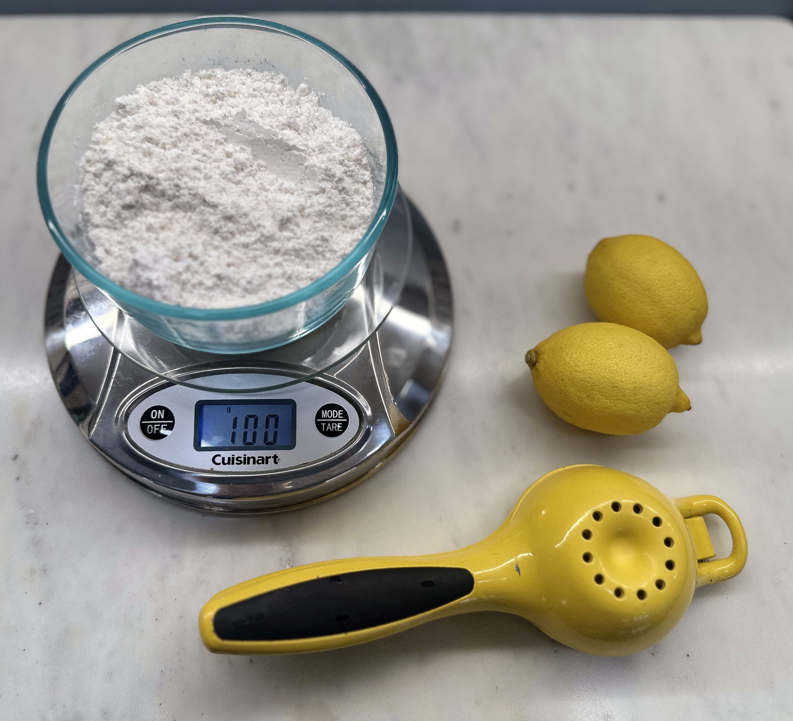 A bowl of powdered sugar sits on a digital kitchen scale, which displays a weight of 100 grams. To the right of the scale are two whole lemons, and in front is a yellow handheld citrus juicer.