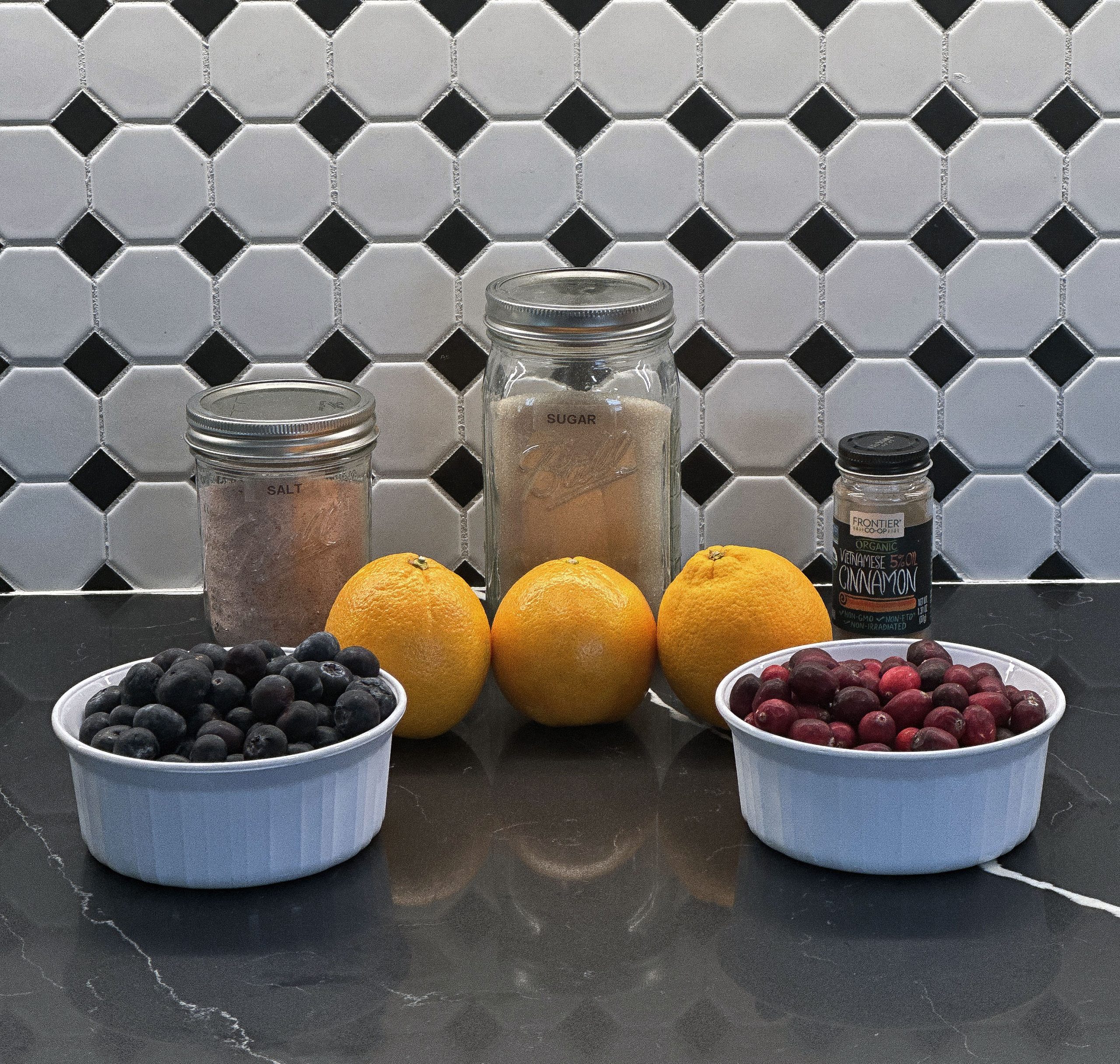 Ingredients for cranberry-blueberry sauce, including fresh blueberries, cranberries, oranges, sugar, salt, and cinnamon, arranged on a black countertop with a tiled background.