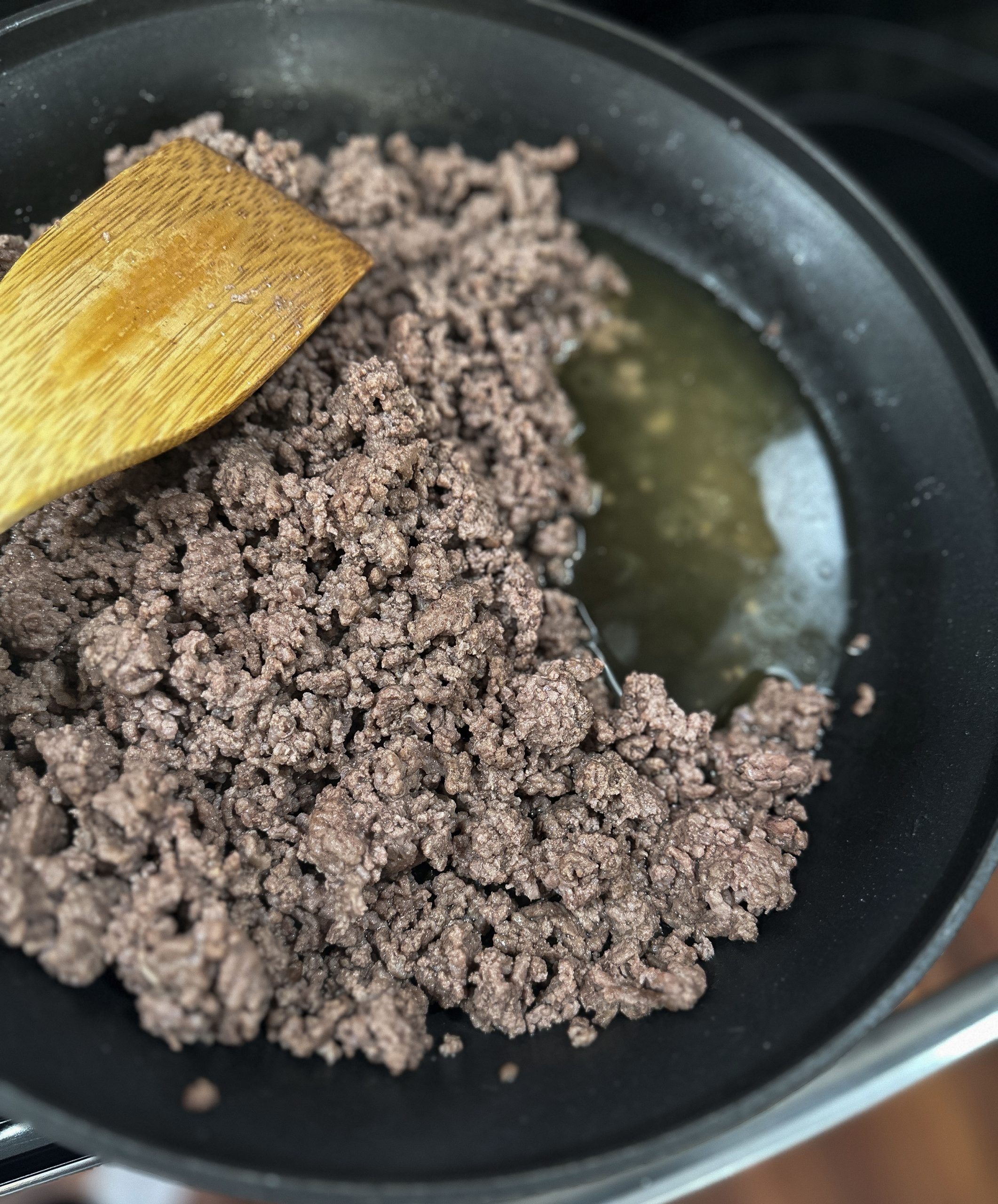 Cooked ground beef in a skillet with rendered fat, being stirred with a wooden spatula.