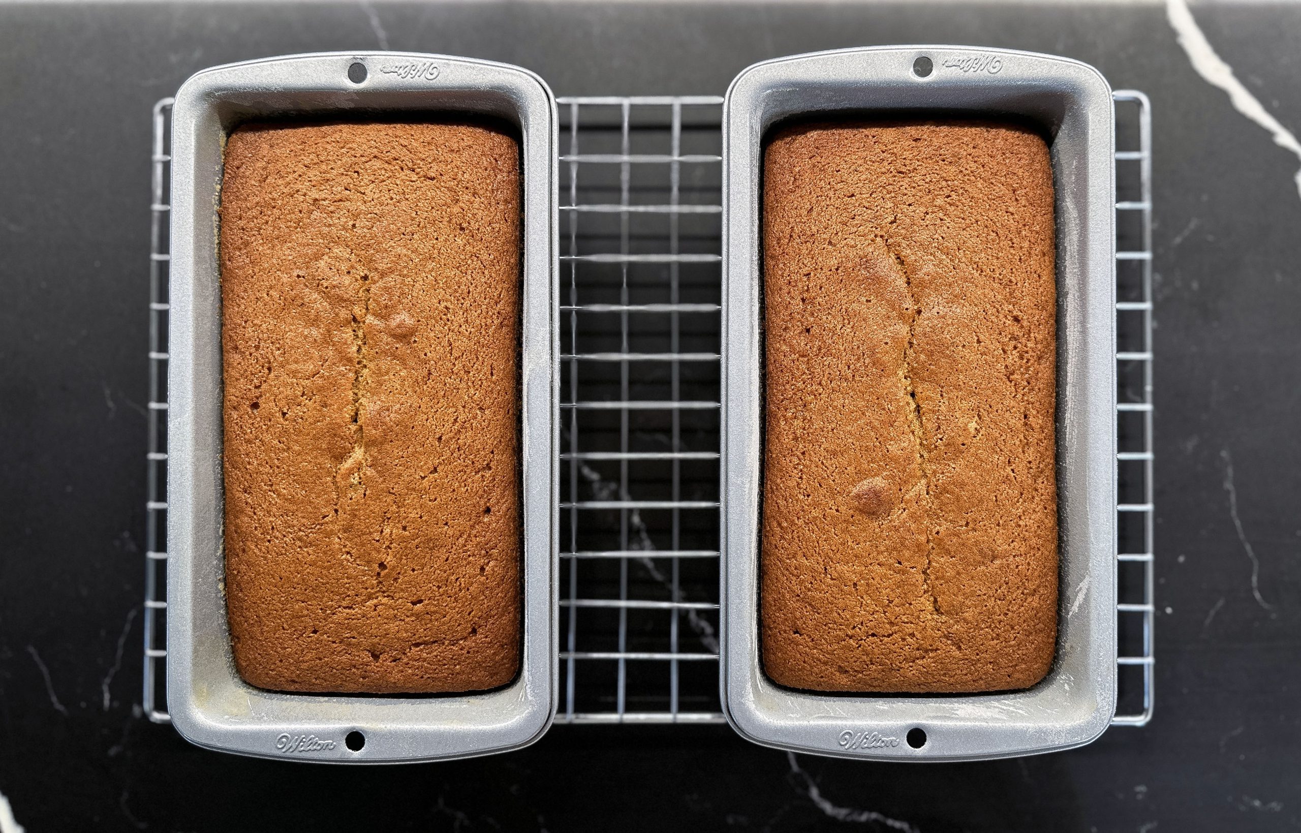 Two freshly baked loaves of pumpkin bread in silver loaf pans, cooling on a wire rack against a dark countertop.
