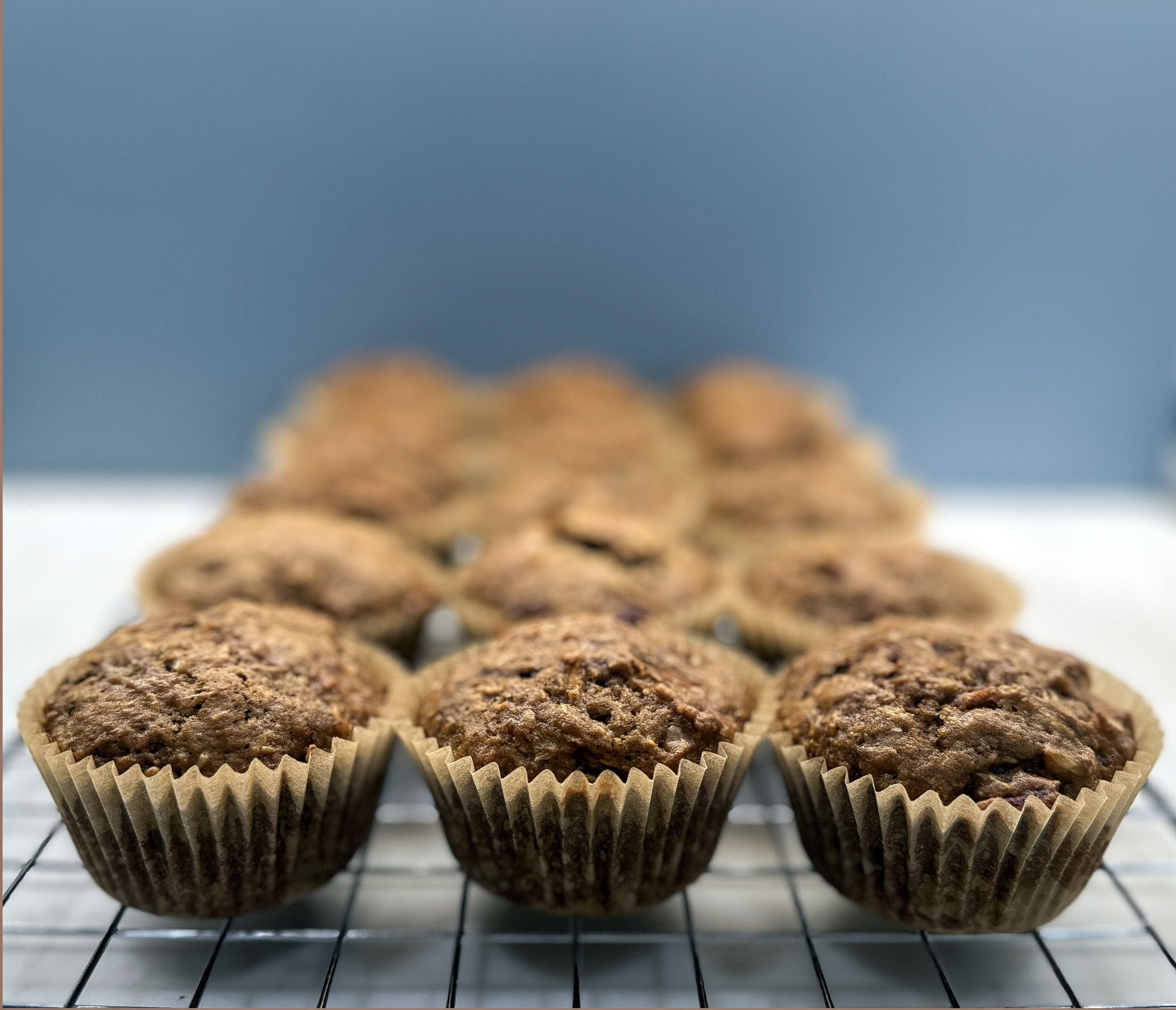 A batch of freshly baked Cranberry Morning Glory Muffins cooling on a wire rack. The muffins are golden brown with slightly cracked tops, showing their hearty texture. They are wrapped in brown paper liners, neatly lined up in rows, with the focus on the muffins in the foreground. 