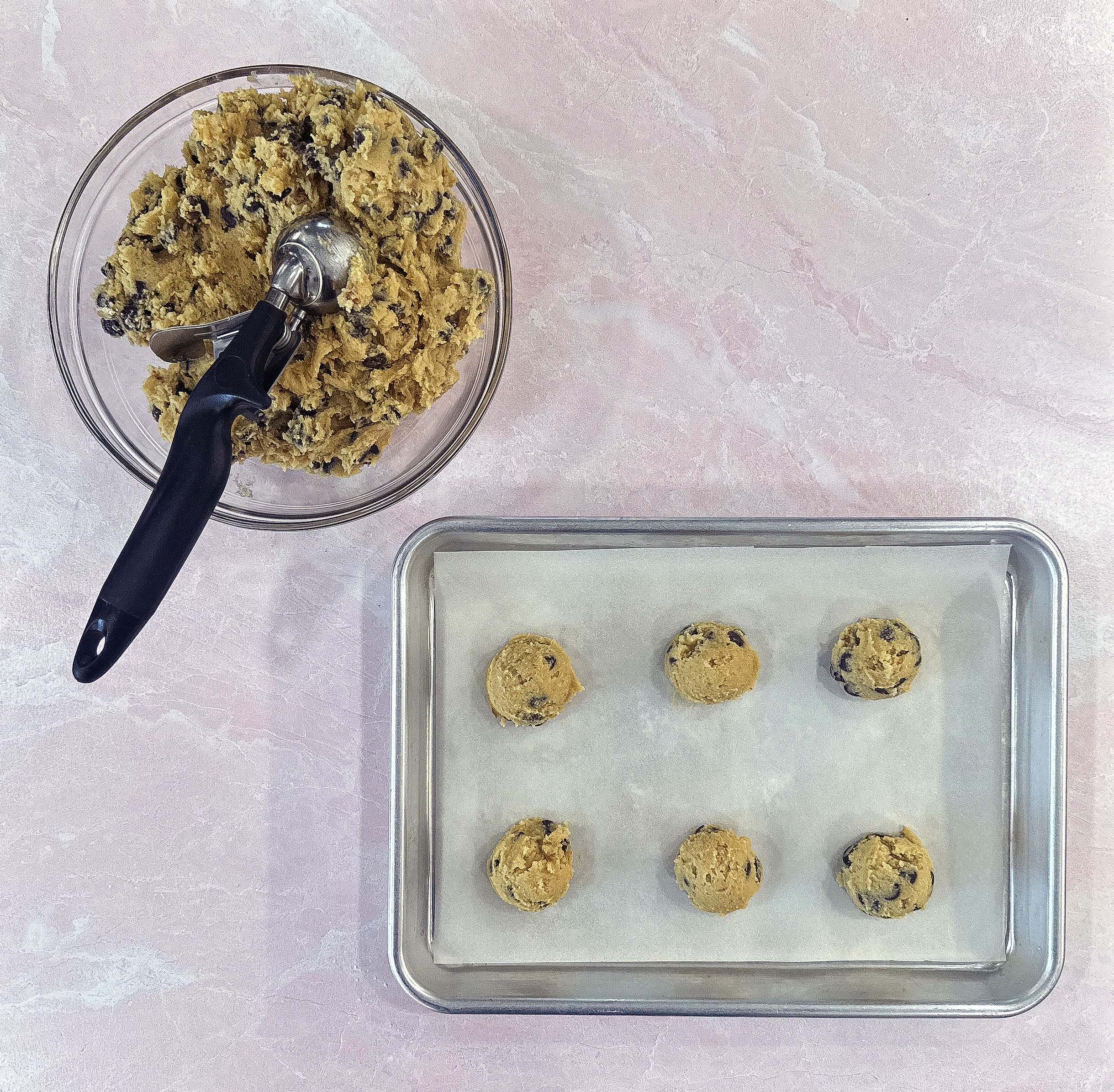 A bowl of chocolate chip cookie dough with a scoop, next to a baking sheet lined with parchment paper holding six scoops of dough.
