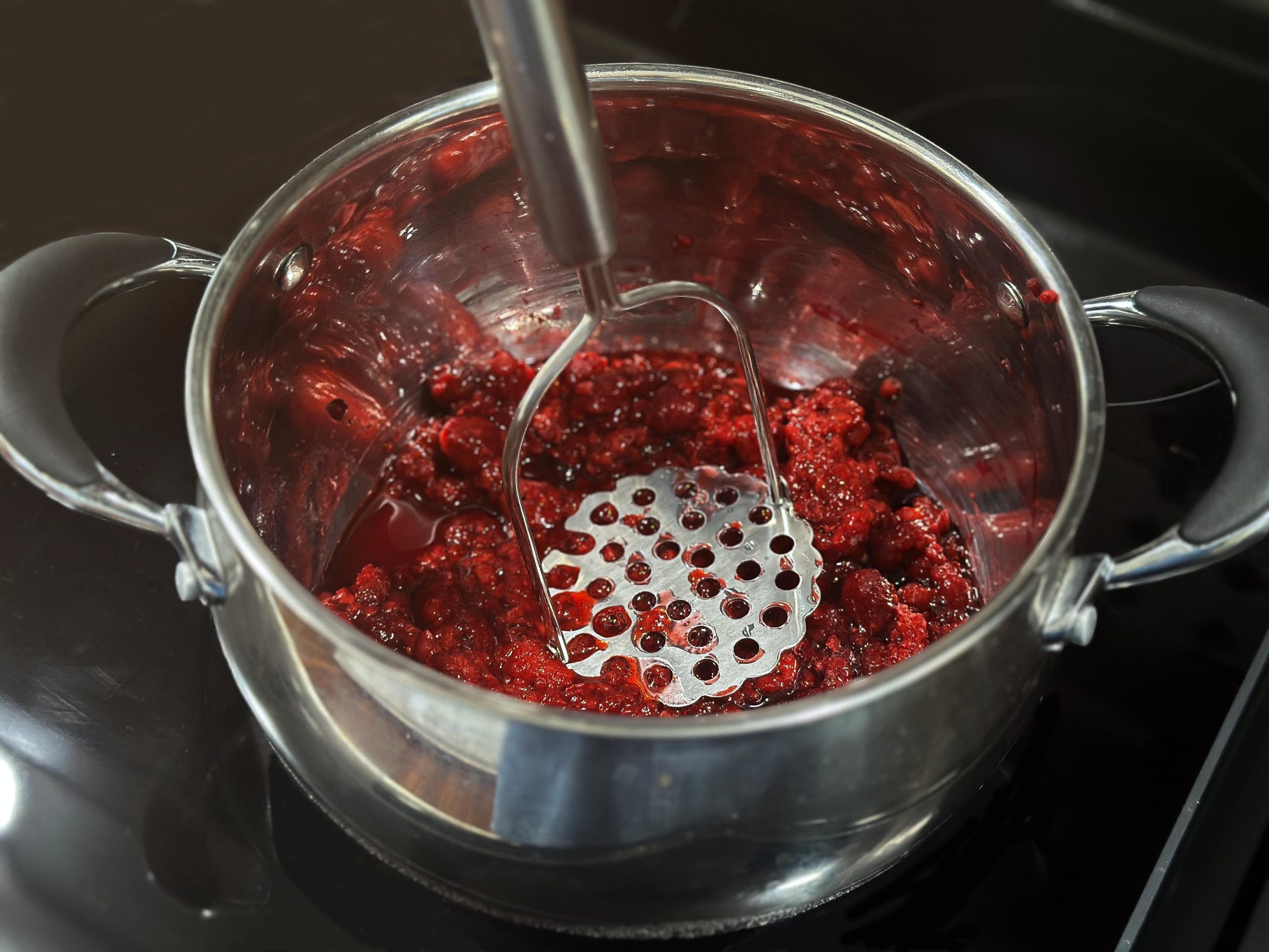 A pot of raspberries being mashed with a metal masher.