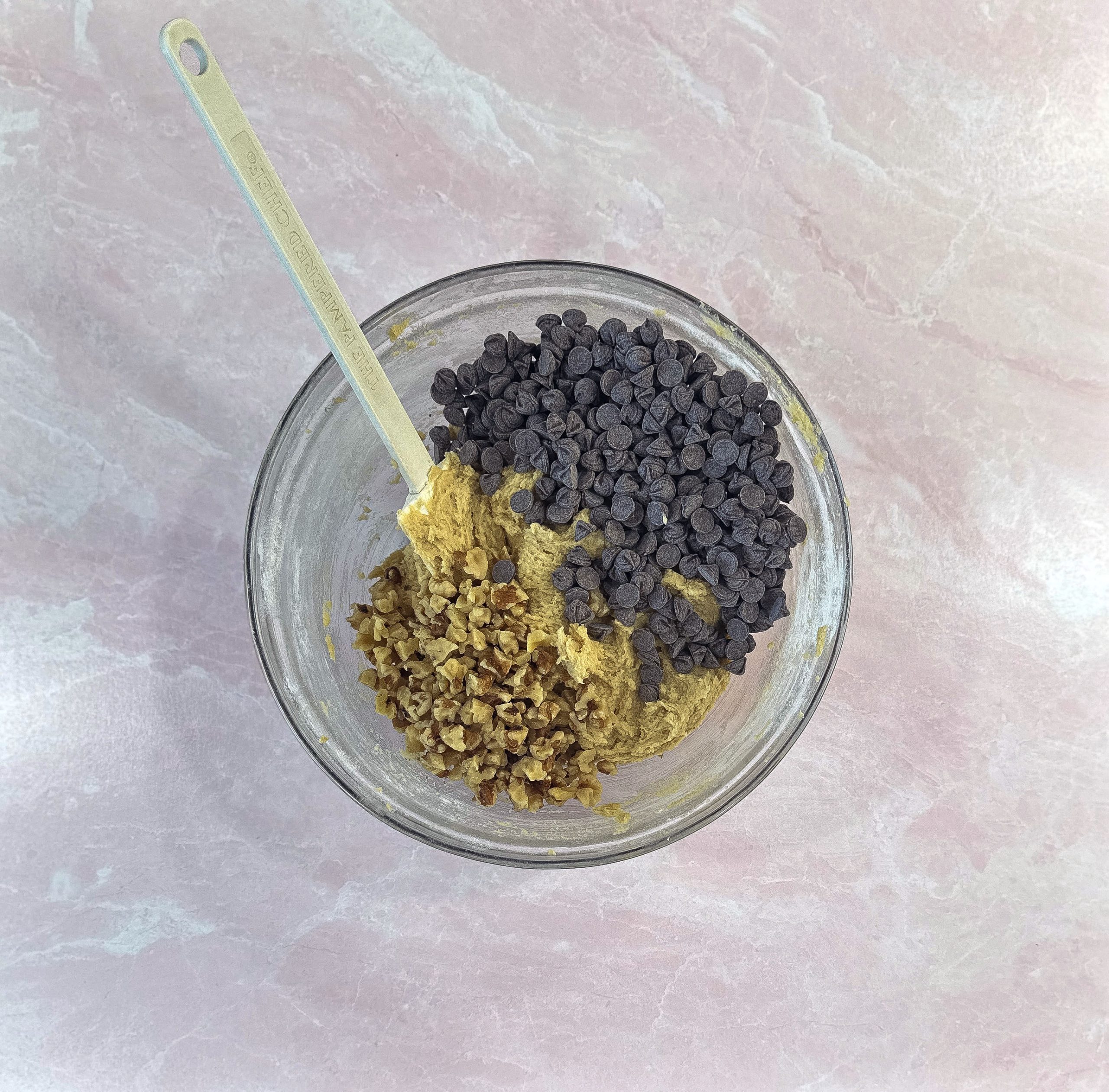 A glass bowl with cookie dough, chocolate chips, and chopped walnuts, partially mixed with a metal spoon on a pink surface.