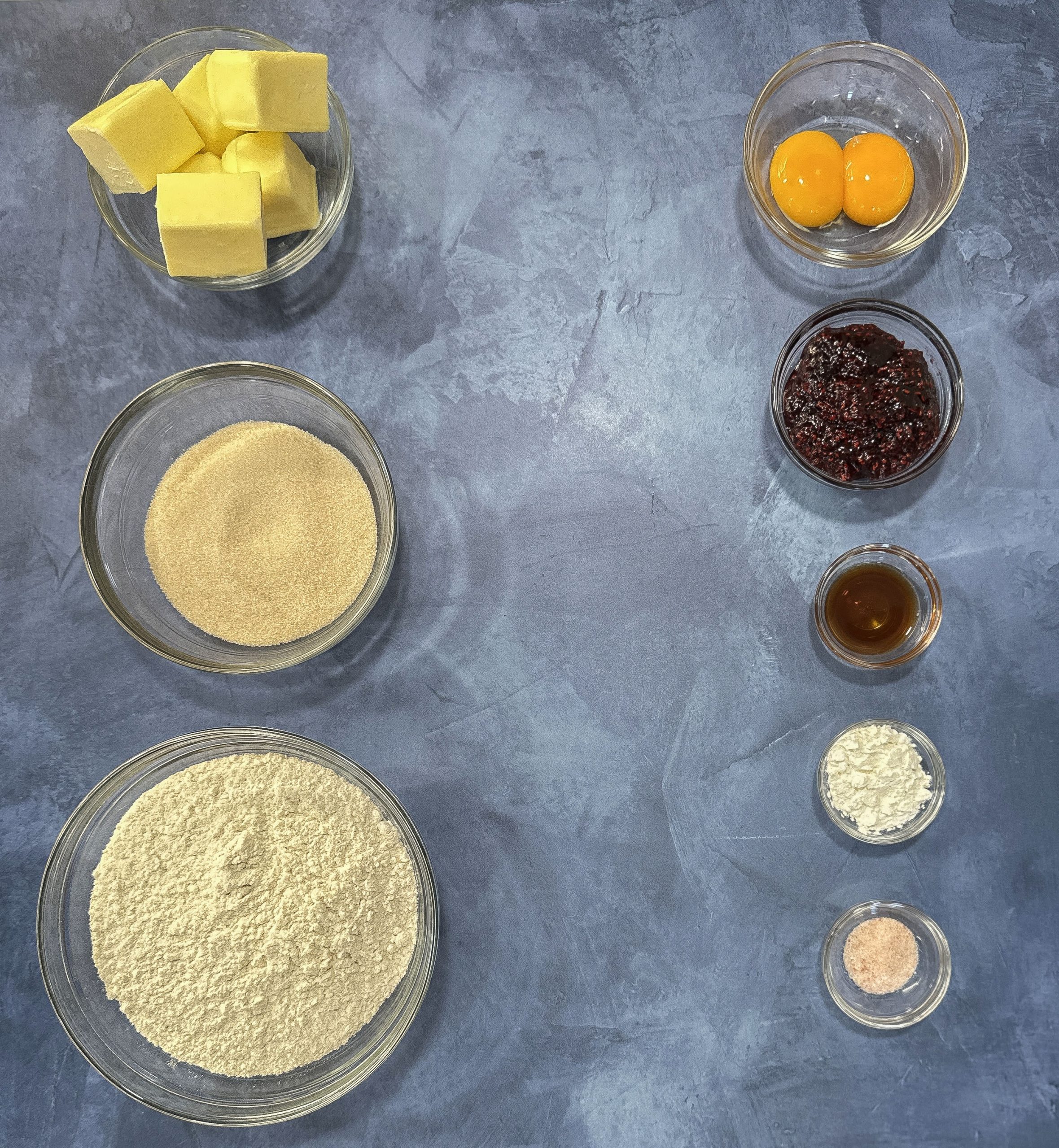 A neatly arranged display of ingredients including butter, sugar, flour, eggs, vanilla extract, and raspberry preserves, ready for baking.
