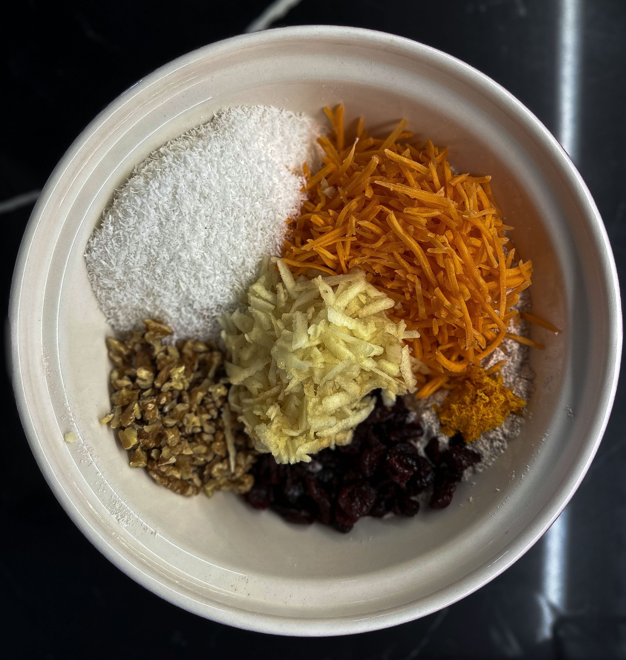 A mixing bowl filled with ingredients for Cranberry Morning Glory Muffins, neatly arranged before mixing. The bowl contains shredded carrots, shredded apple, chopped walnuts, dried cranberries, orange zest, shredded coconut, and flour. Each ingredient is placed separately in the bowl, showcasing the variety of textures and colors. The bright orange carrots and zest contrast with the cranberries and walnuts, giving a vibrant preview of the muffin mix.