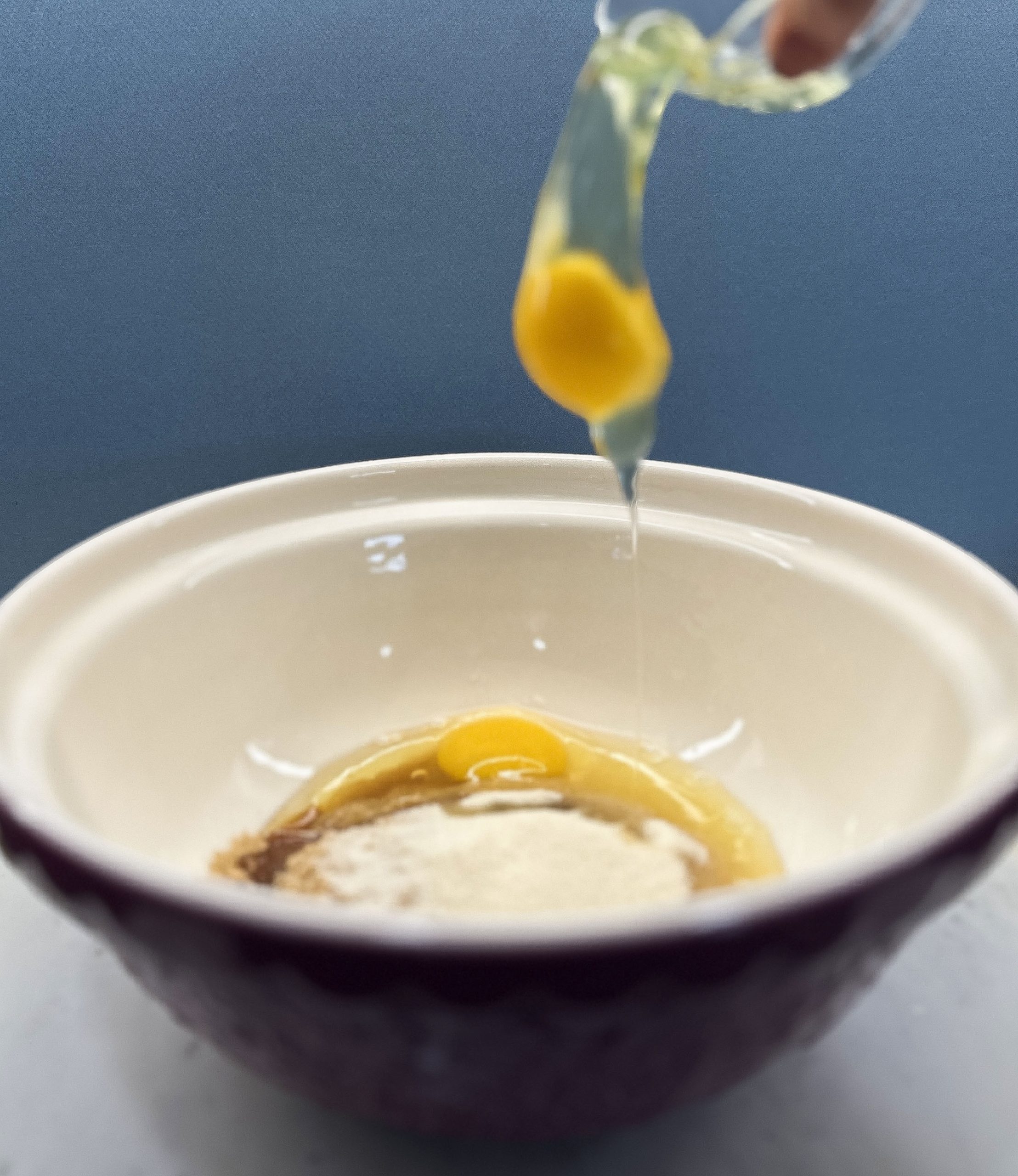 A close-up of an egg dropped into a mixing bowl that contains sugar and other ingredients, preparing the wet mixture for Double Chocolate Zucchini Bread. The action shot captures the egg mid-air as it drops into the bowl.