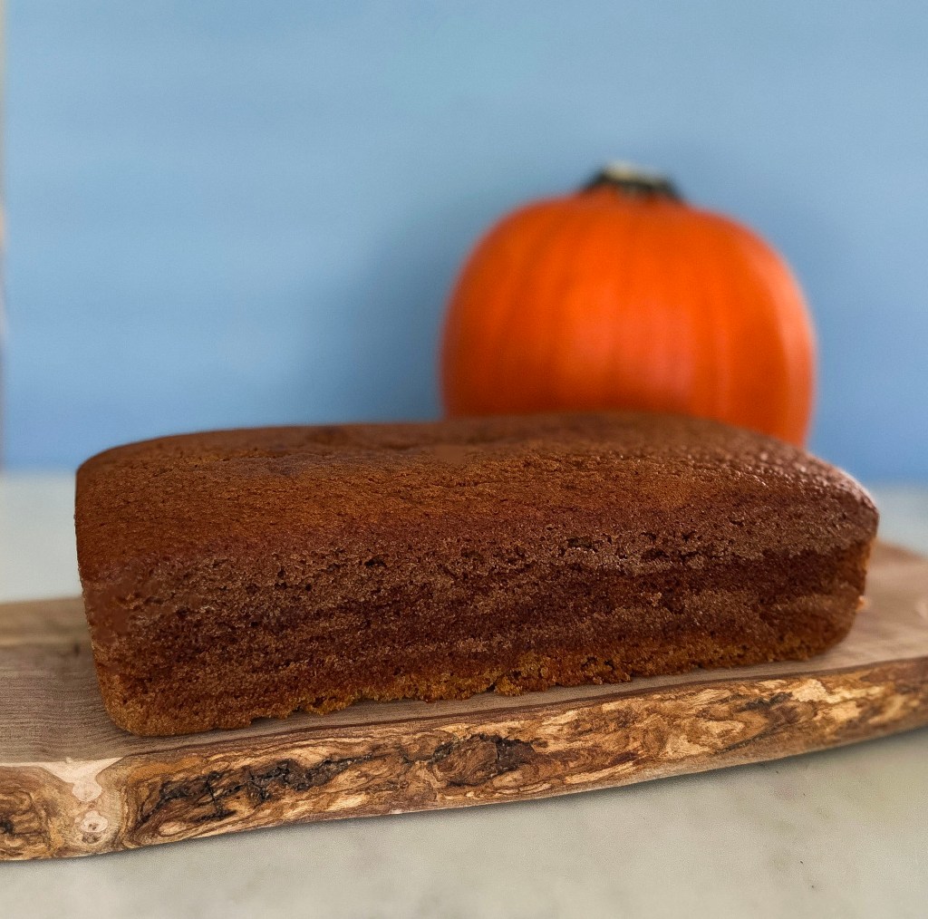 A loaf of pumpkin bread resting on a rustic wooden board, with a deep brown top and a soft crumb, set against a backdrop of an orange pumpkin.