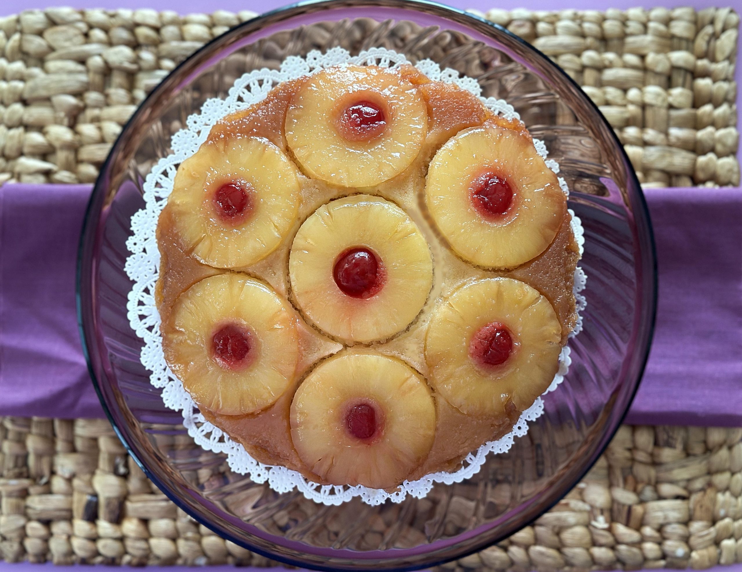 Pineapple Upside Down Cake with seven pineapple rings and cherries in the center, placed on a glass plate with a purple cloth and wicker mat.