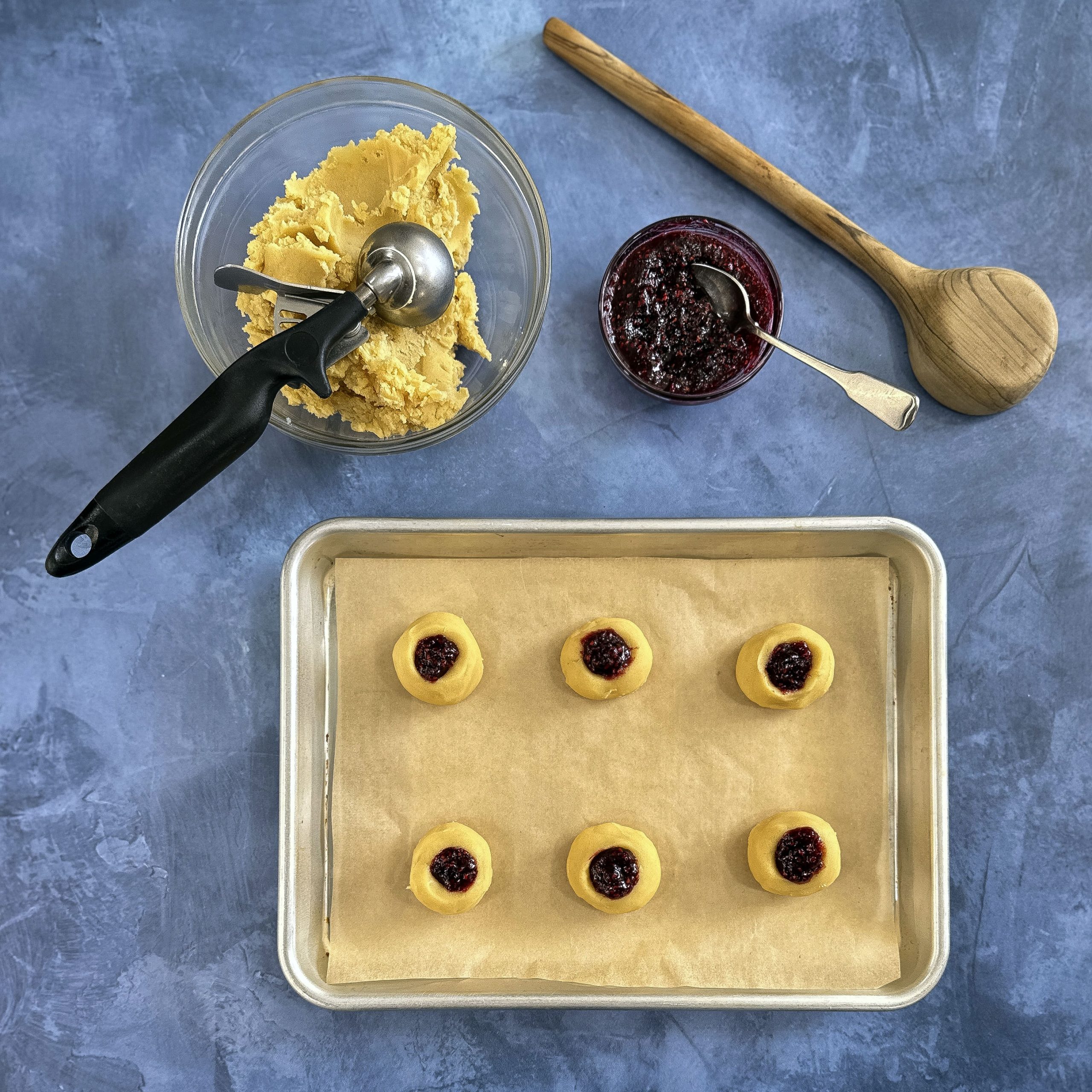 A baking sheet filled with assembled raspberry thumbprint cookies, each with a neatly pressed indentation and a dollop of warm raspberry preserves before baking.