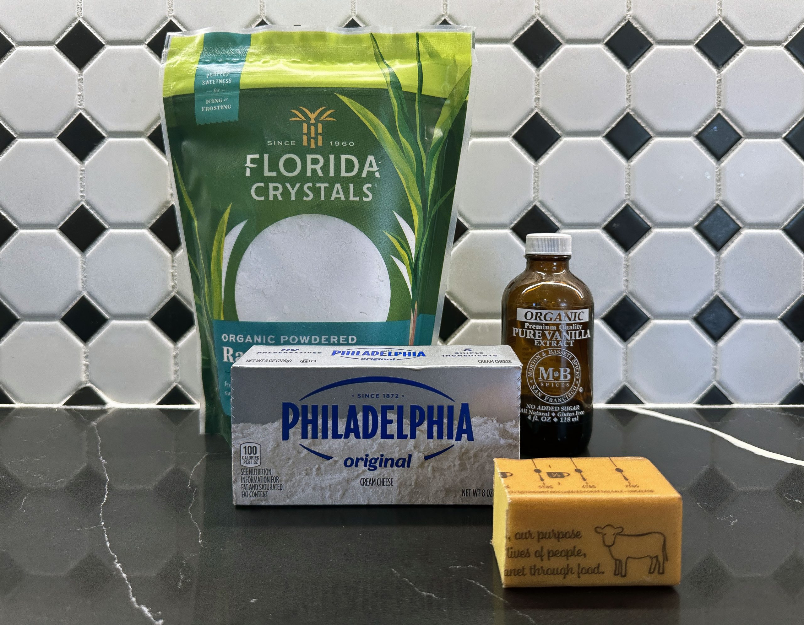 A selection of buttercream filling ingredients, including powdered sugar, cream cheese, butter, and vanilla extract, displayed on a black countertop with a tiled backsplash.