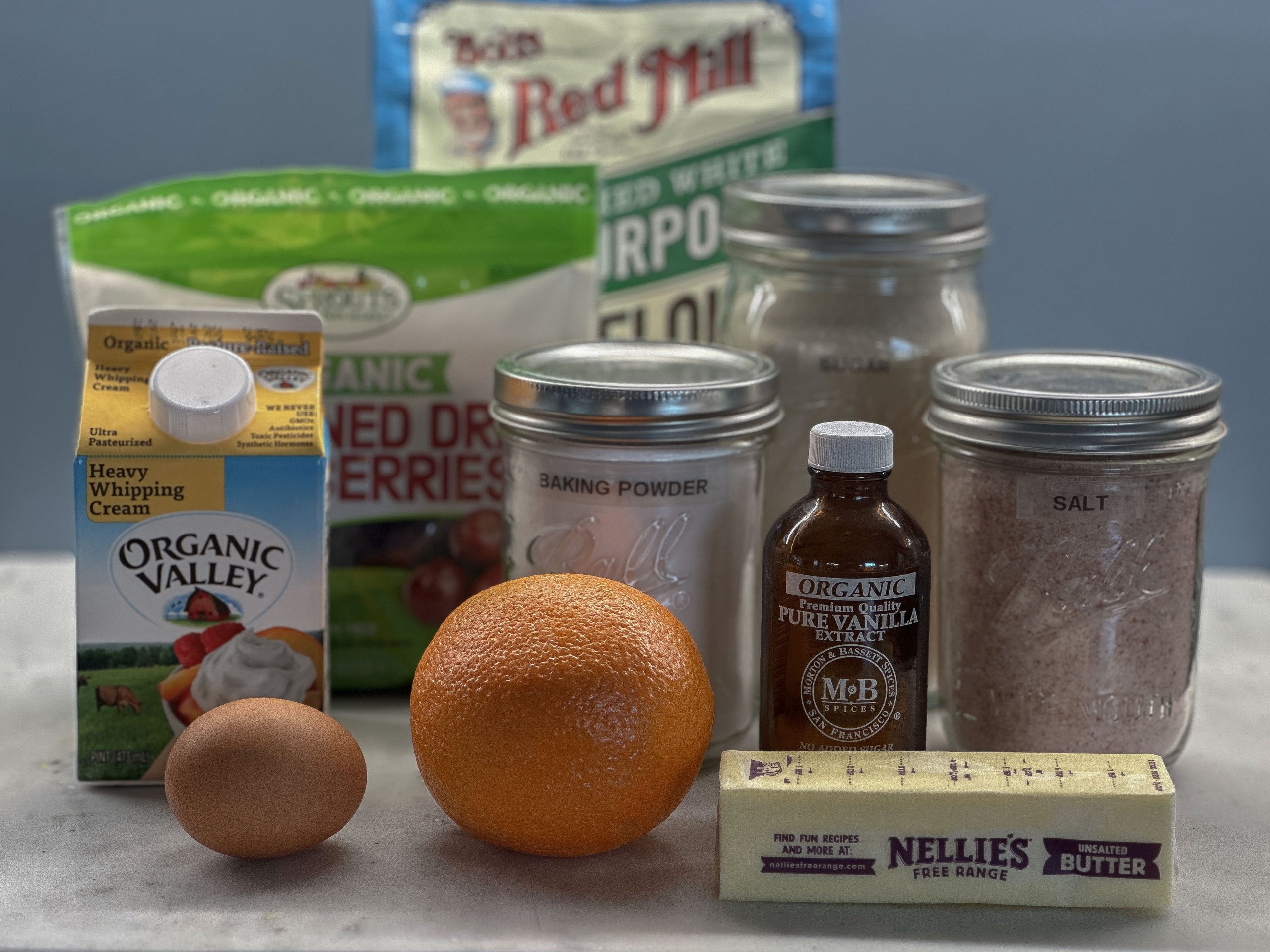 A collection of ingredients for cranberry orange scones is arranged on a countertop. The ingredients include a carton of Organic Valley heavy whipping cream, an egg, an orange, a stick of Nellie’s Free Range unsalted butter, a bottle of organic vanilla extract, and jars labeled with baking powder, salt, and sugar. In the background, a bag of Bob’s Red Mill all-purpose flour and a package of dried cranberries are also visible. These are the essential components for preparing a batch of cranberry orange scones.