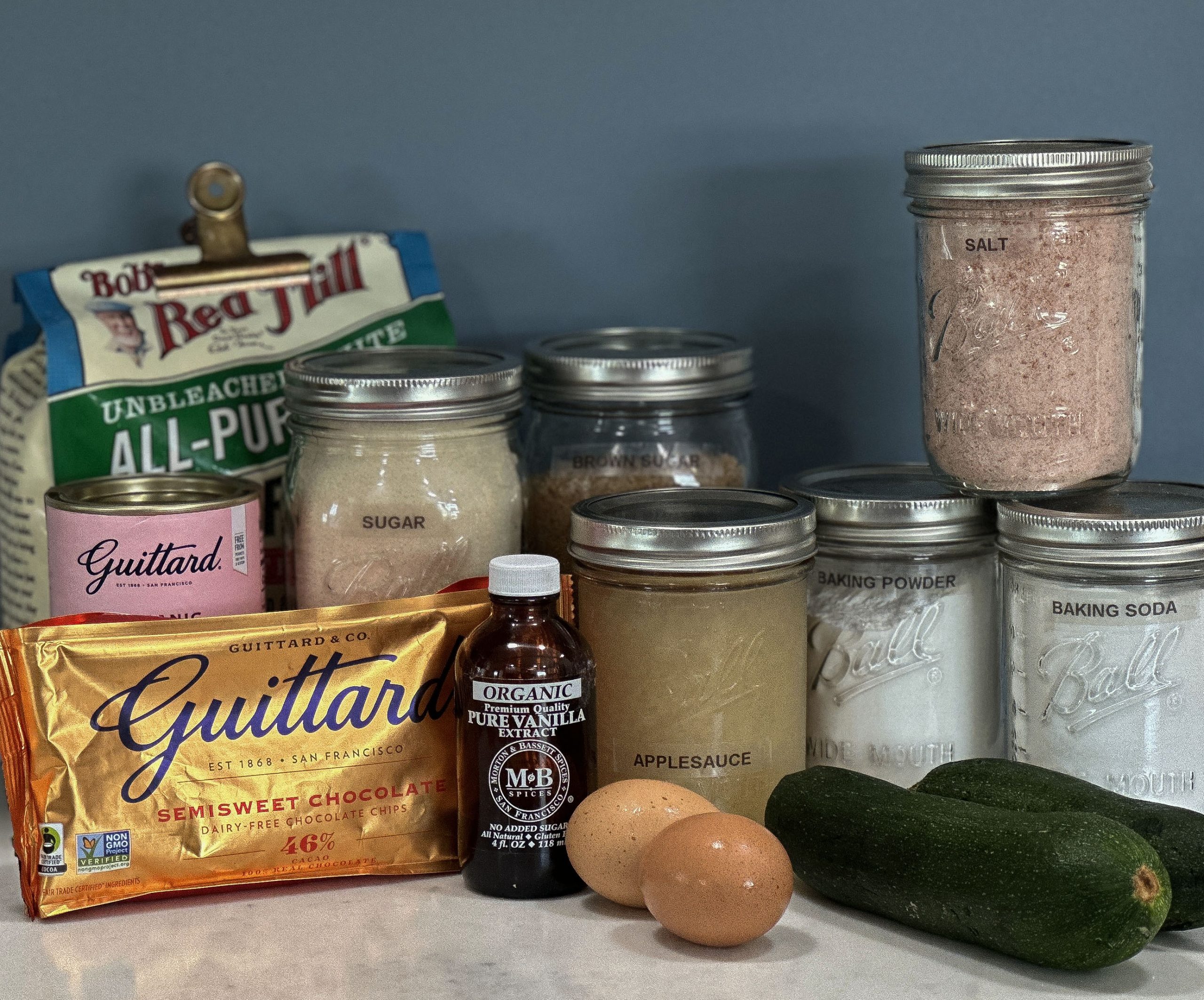Ingredients for Double Chocolate Zucchini Bread neatly arranged on a countertop. The image includes all-purpose flour, sugar, brown sugar, salt, baking powder, baking soda, applesauce, vanilla extract, chocolate chips, eggs, and fresh zucchinis. The ingredients are a mix of branded items, such as Guittard chocolate and vanilla extract, and staple pantry items stored in mason jars.