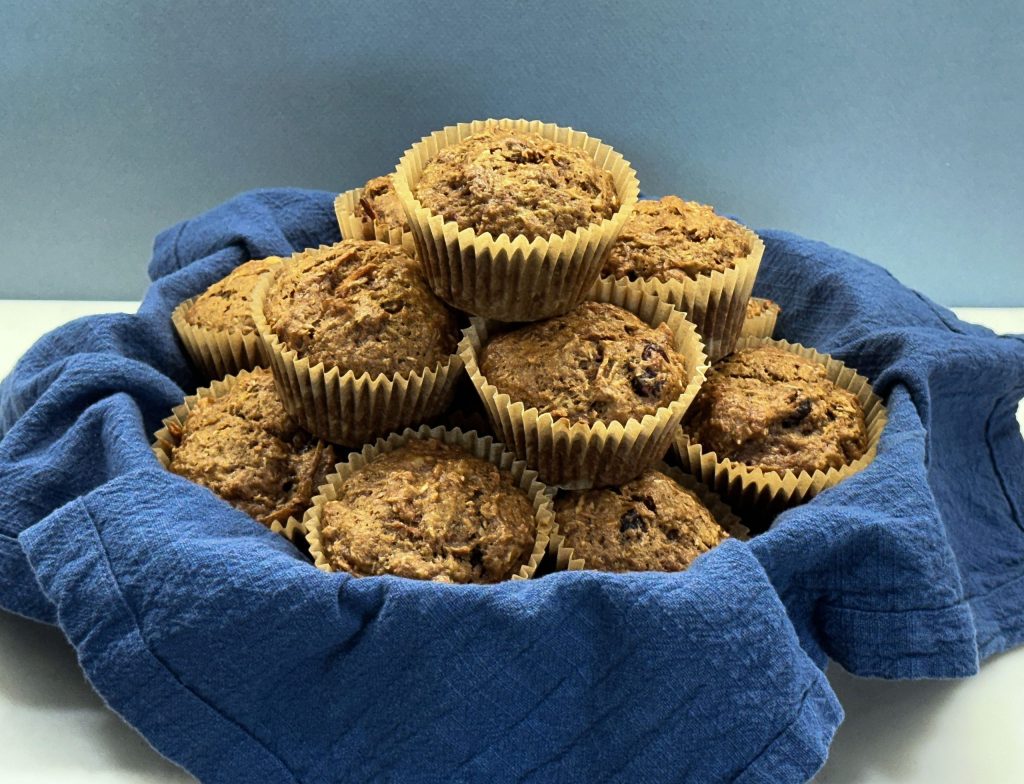 A basket filled with golden-brown Cranberry Morning Glory Muffins wrapped in brown paper liners. The muffins are stacked in a cozy blue cloth, which lines the basket, creating a warm and inviting presentation. The muffins appear moist with visible bits of cranberries, and their tops are slightly cracked, showing the hearty texture of ingredients like carrots and oats. The background is a simple, muted blue-gray, which helps the muffins stand out as the centerpiece.