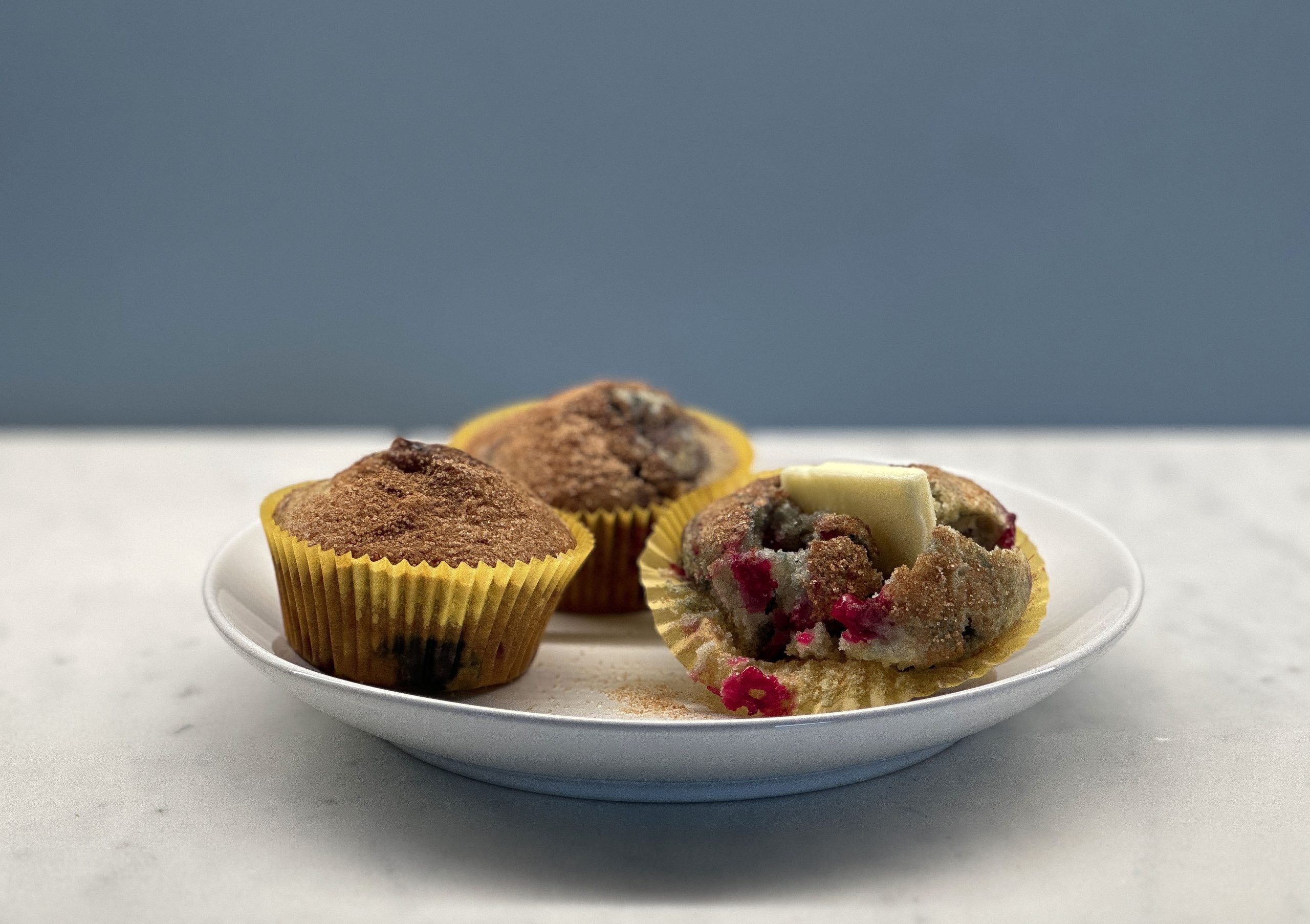 Three berry muffins on a white plate, with one muffin partially unwrapped and topped with a pat of butter.