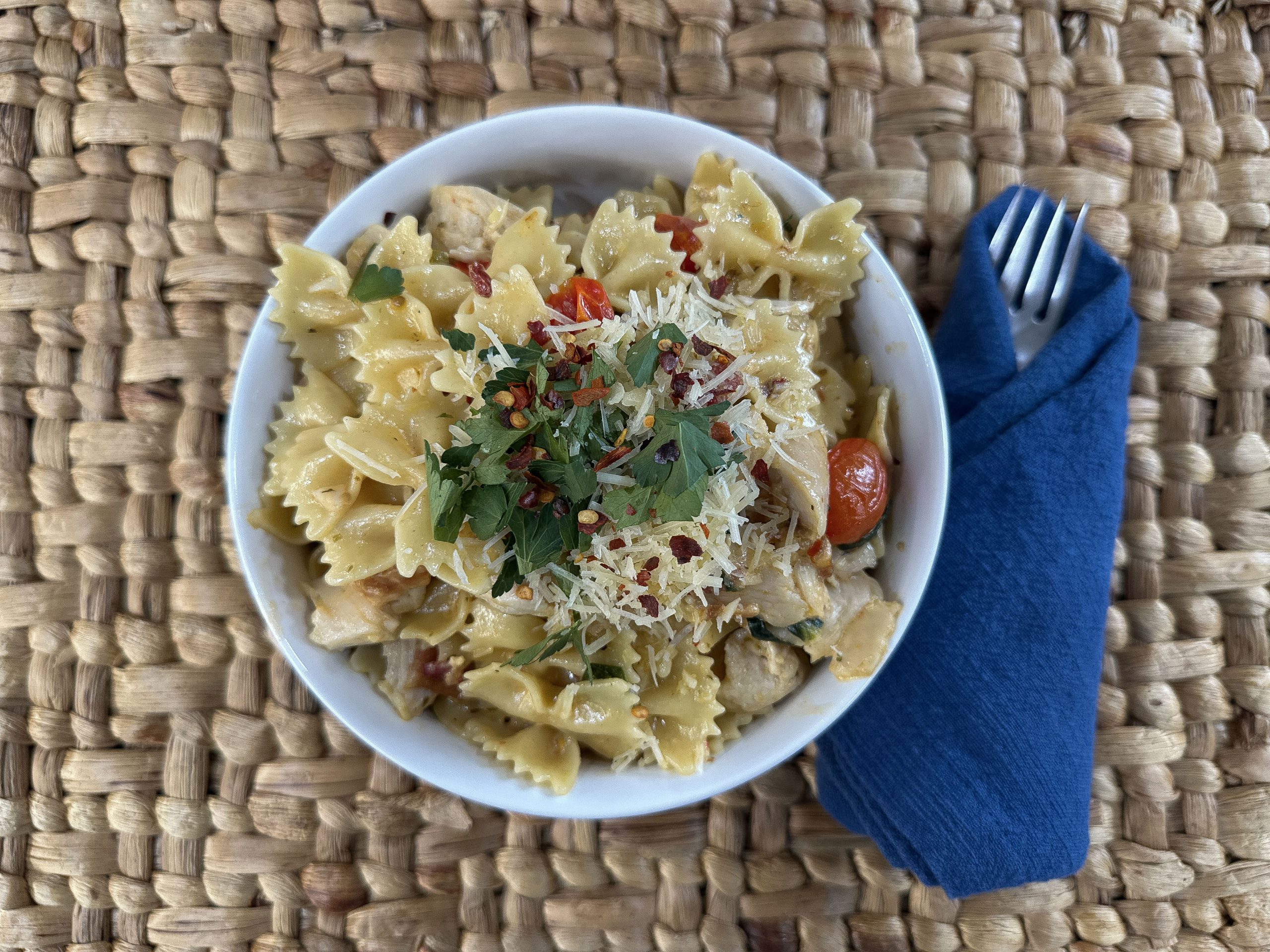 Overhead view of the finished creamy pasta dish served in a white bowl on a woven placemat with a blue napkin and fork.
