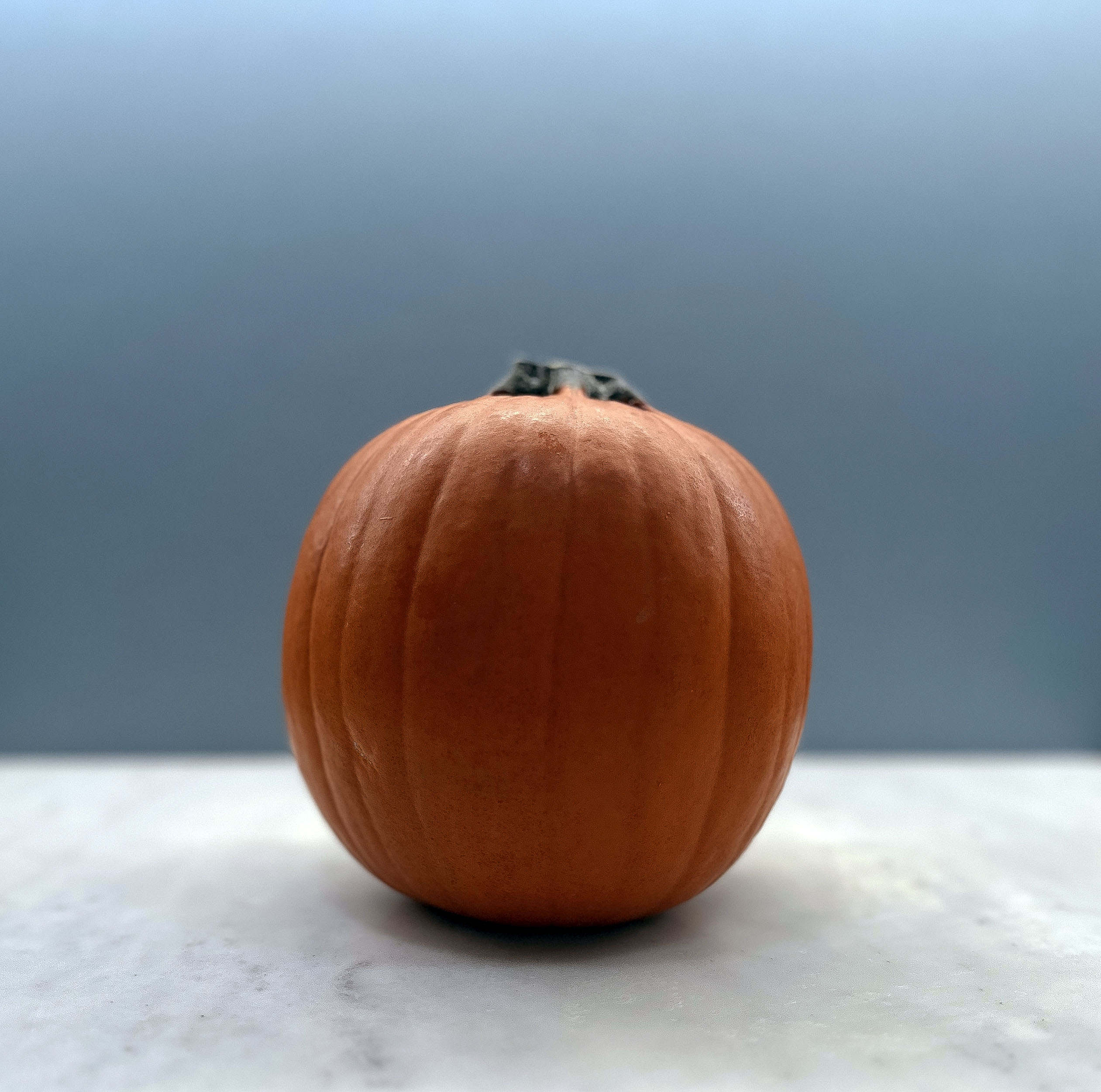This image features a small, round orange pumpkin with a dark green stem on a light surface against a soft blue background.