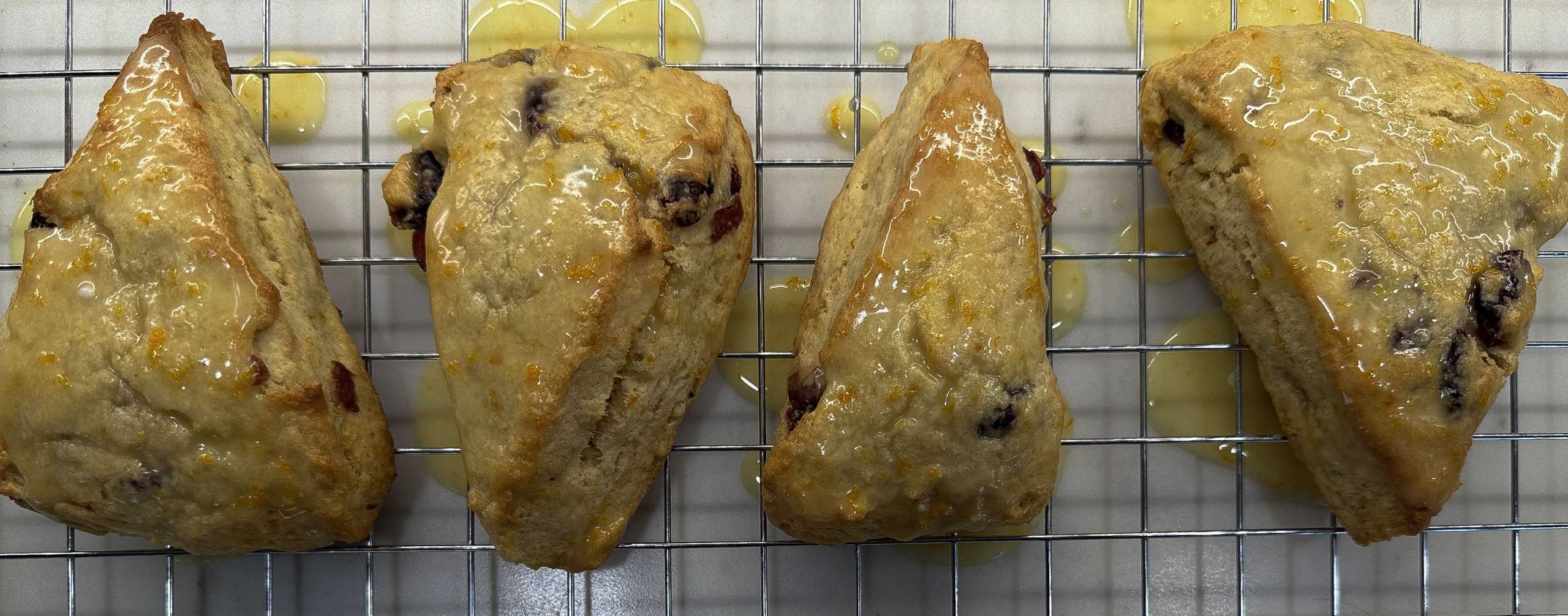 Four golden-brown cranberry orange scones sit on a wire cooling rack. Each scone is topped with a shiny drizzle of orange glaze, which has dripped slightly onto the rack below. The scones are triangular in shape with visible bits of cranberry peeking through the dough, offering a delicious balance of tart and sweet flavors.