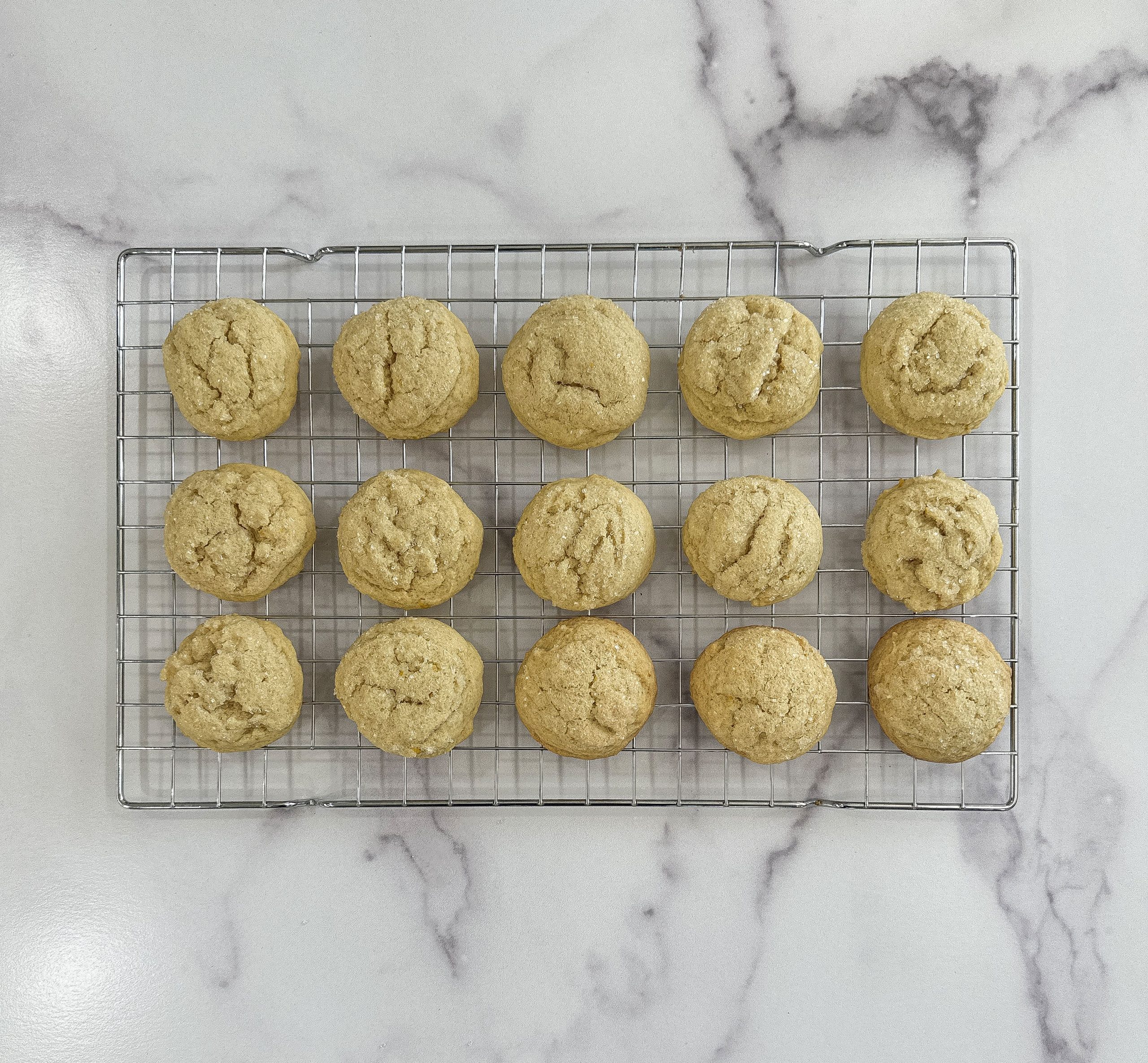 Freshly baked lemon sugar cookies cooling on a wire rack, with a golden, slightly cracked surface on a marble countertop.