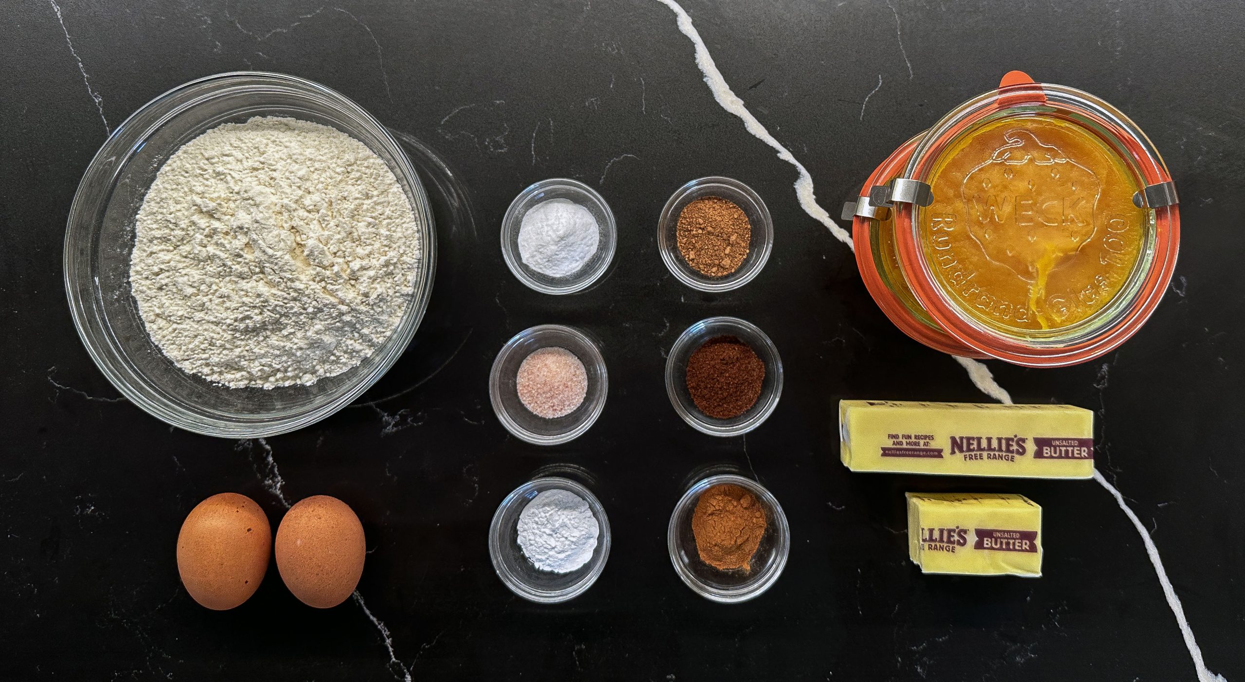 A top-down view of ingredients for pumpkin bread, including flour, eggs, butter, pumpkin puree, and various spices, neatly arranged on a black countertop.
