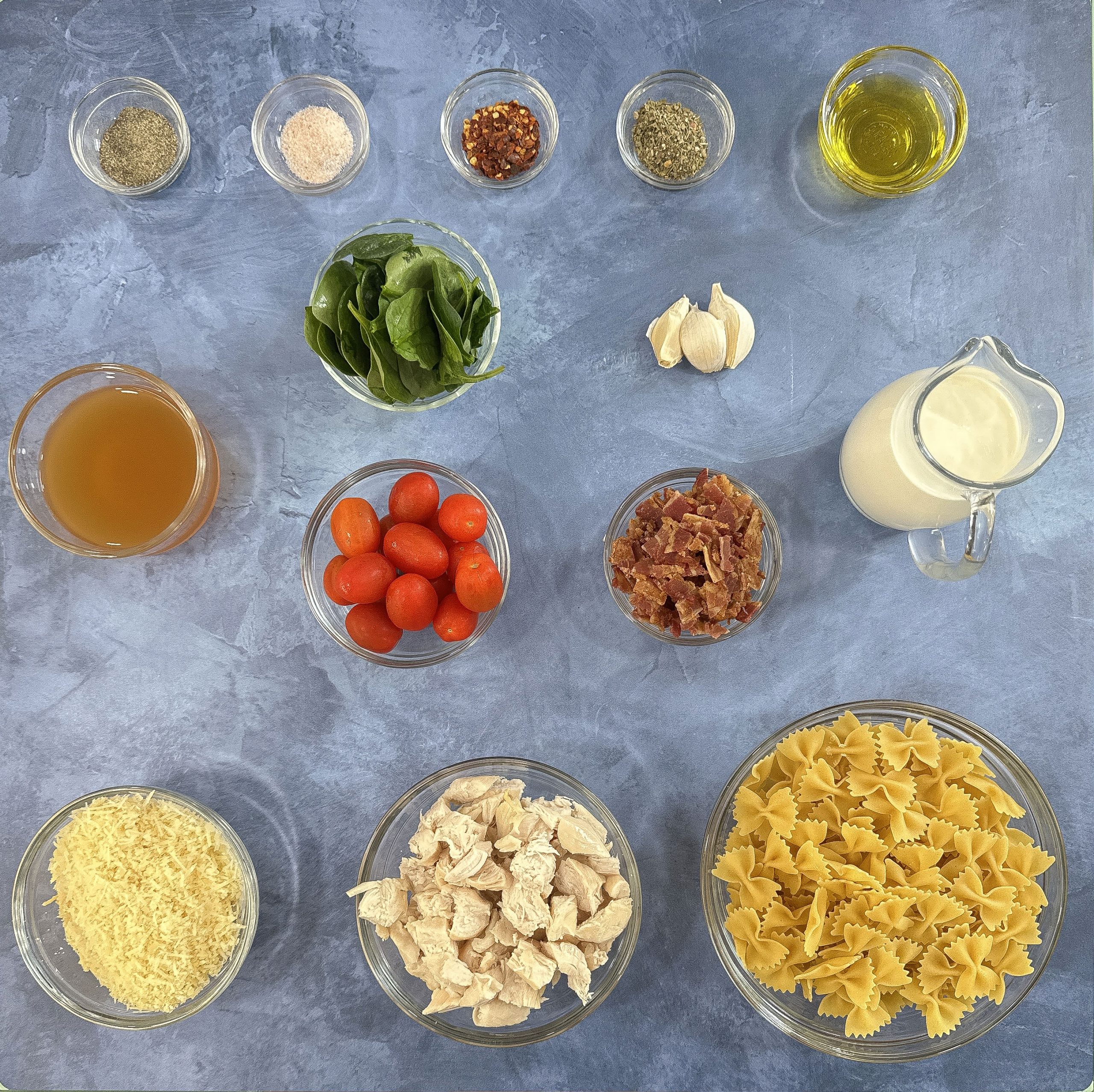 Overhead shot of measured ingredients for creamy pasta with chicken and bacon, including pasta, cherry tomatoes, spinach, cooked chicken, bacon, garlic, cream, broth, and seasonings.
