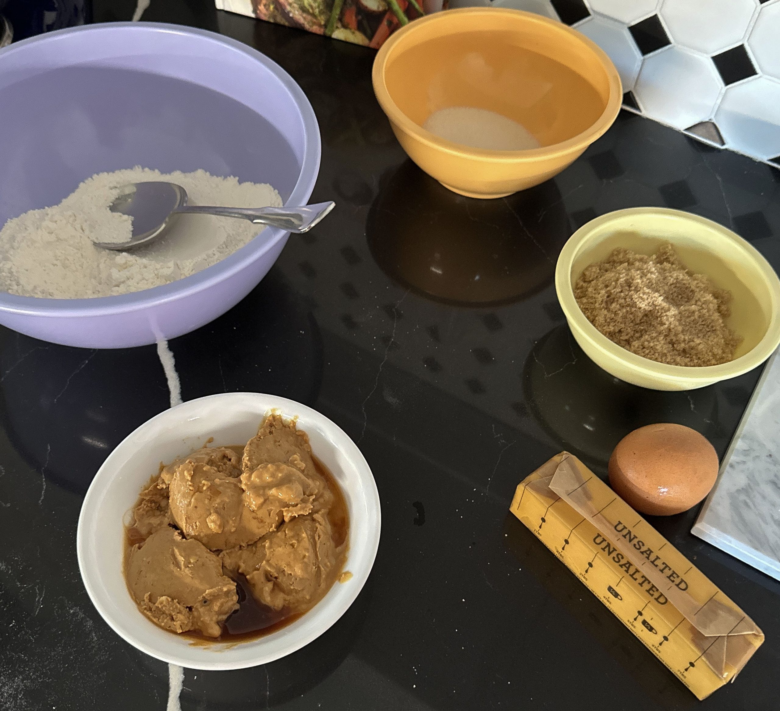 Ingredients for peanut butter cookies arranged on a black countertop, including bowls of flour, sugar, brown sugar, a dish with peanut butter and vanilla extract, a stick of unsalted butter, and a single egg.