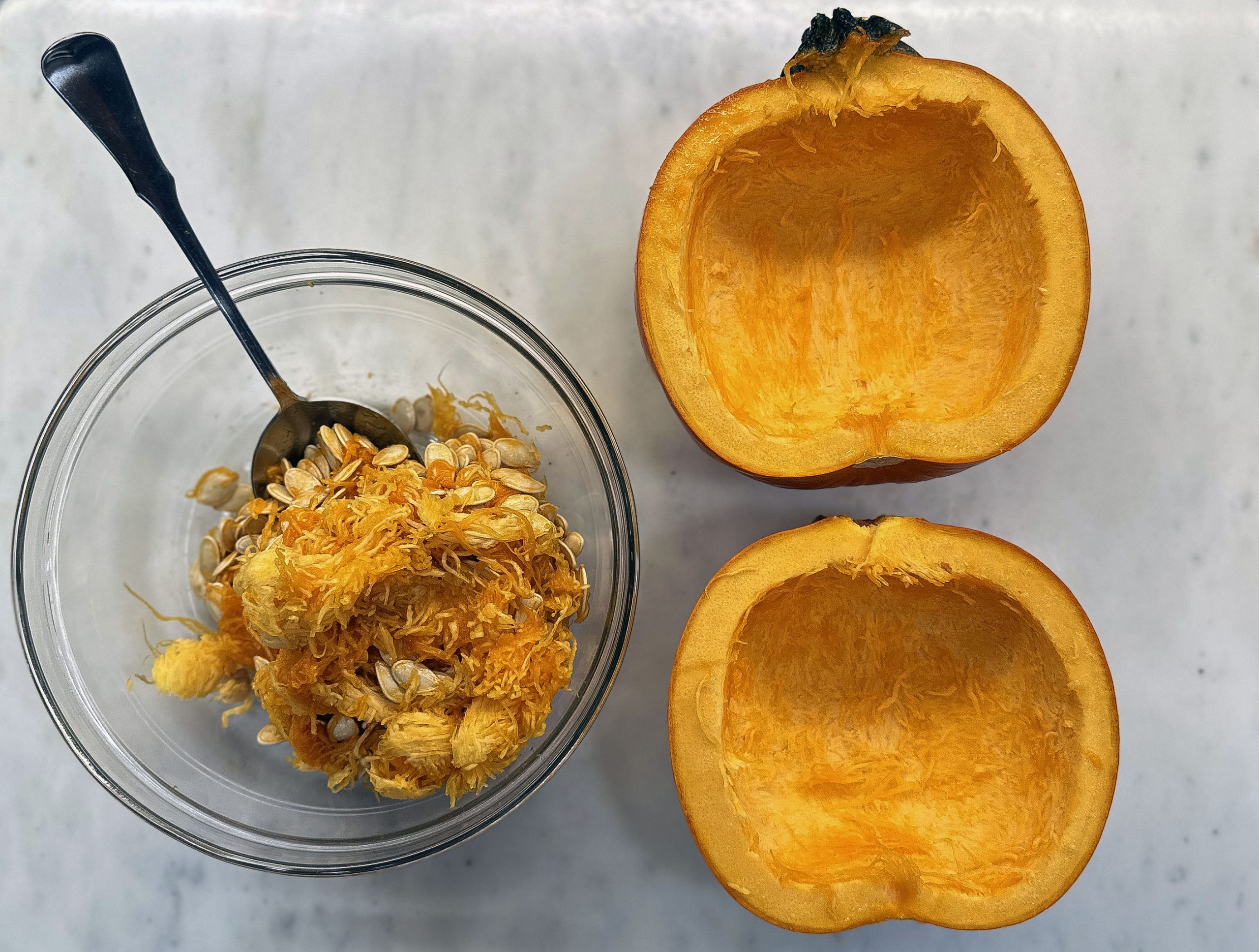This image shows a halved pumpkin with its seeds and pulp scooped into a glass bowl on the left, alongside a spoon, all set on a light-colored surface.