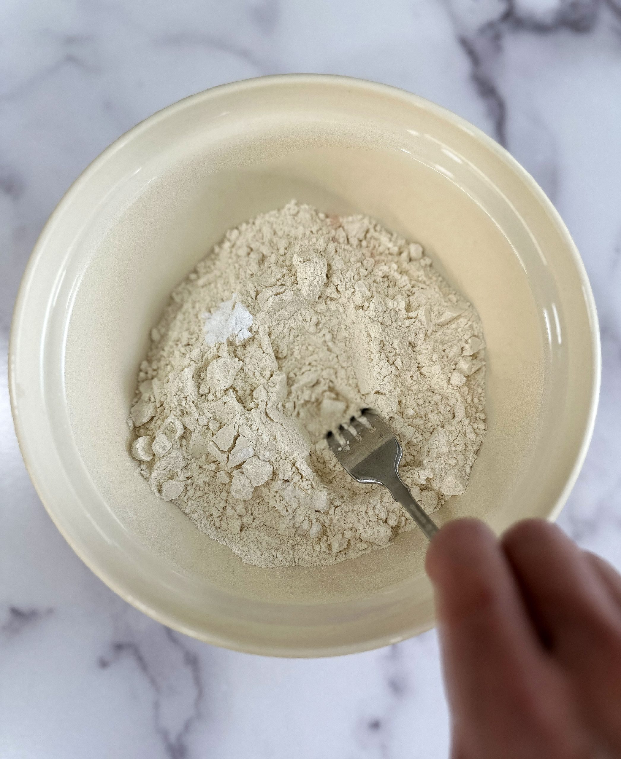 A hand using a fork to mix dry ingredients, including flour, baking soda, and baking powder, in a beige mixing bowl on a marble countertop.