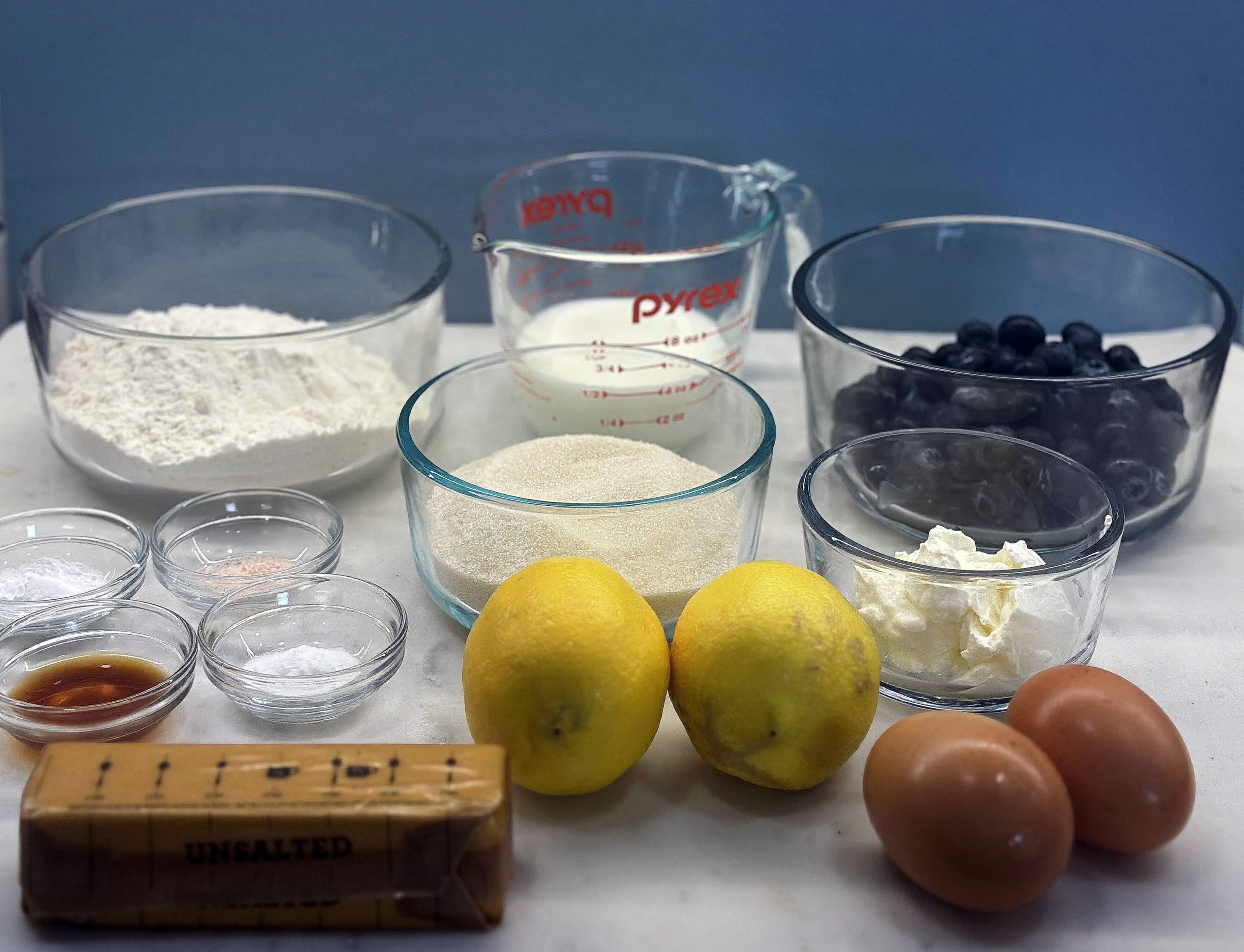 The image shows ingredients for this recipe. The items shown include:
a bowl of flour, measuring cup of milk, a bowl of blueberries, two lemons, two eggs, a stick of unsalted butter, a bowl of sugar, a bowl with a dollop of sour cream cheese, several small bowls containing baking powder, baking soda, salt, and vanilla extract
