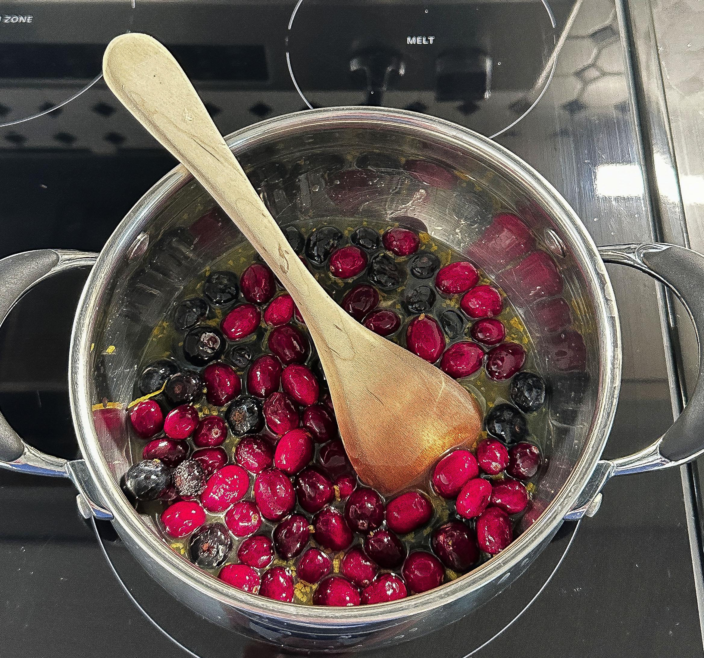 Cranberries and blueberries cooking in a saucepan with a wooden spoon on a stovetop.