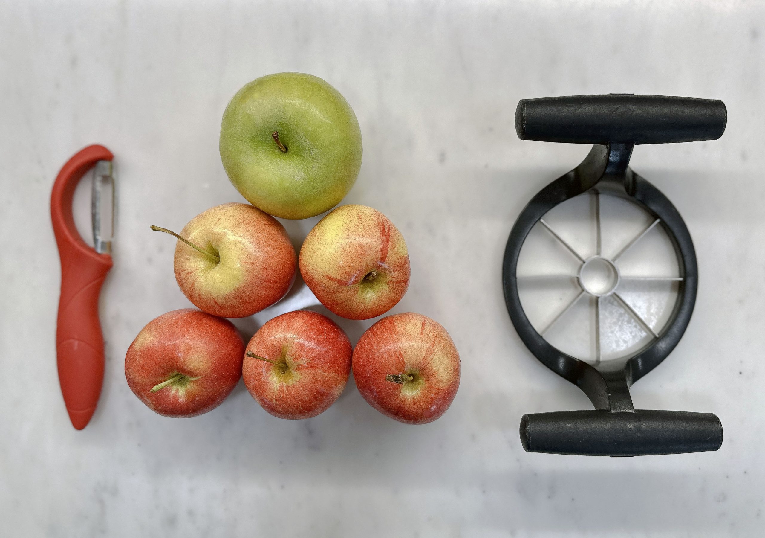 An overhead view of a black apple slicer, a group of apples including one green and several red, and an orange vegetable peeler arranged on a white marble surface.