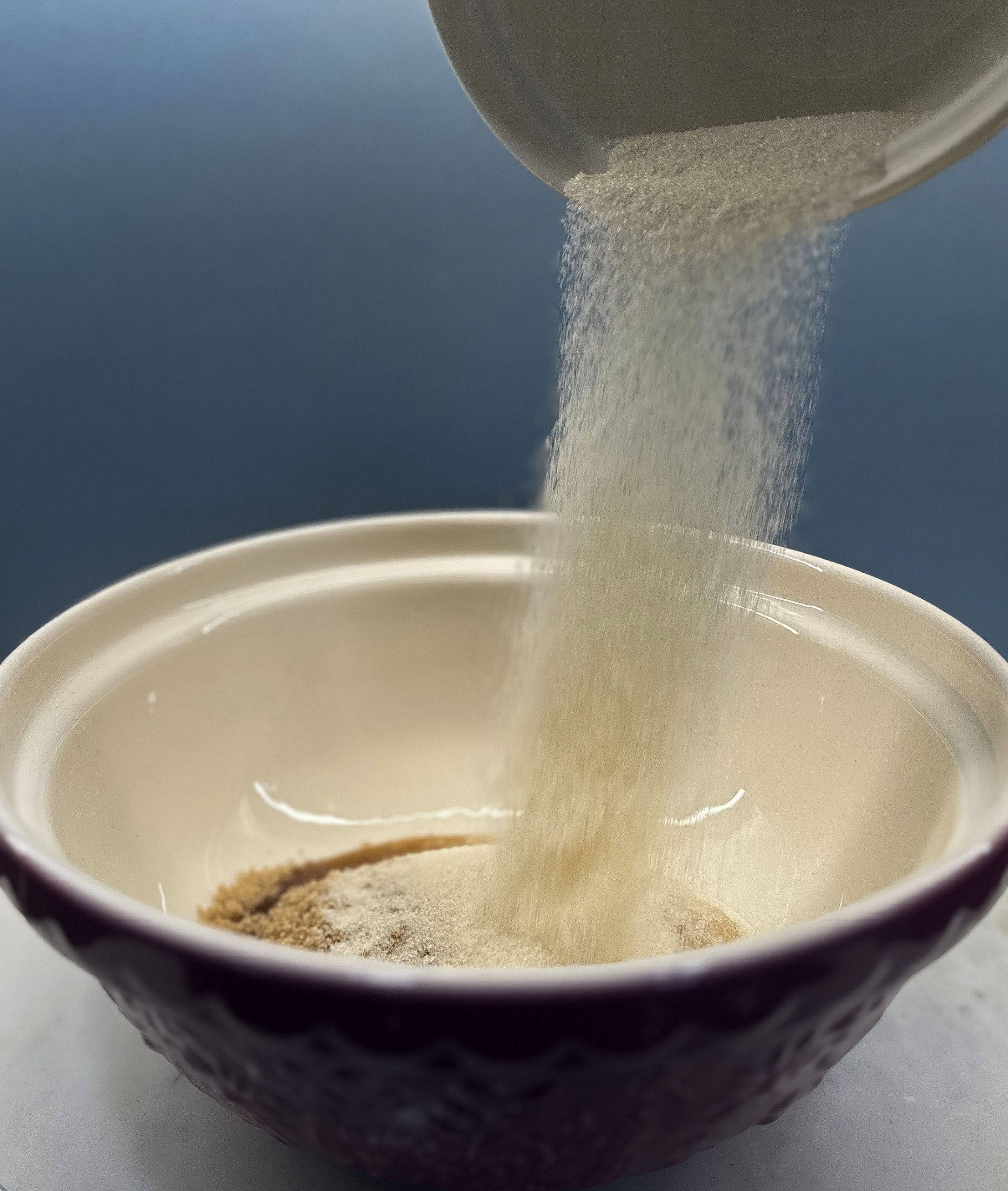 A close-up of sugar being poured into a mixing bowl containing other dry ingredients, in preparation for making Double Chocolate Zucchini Bread. The sugar is caught mid-pour, creating a cascade into the bowl.