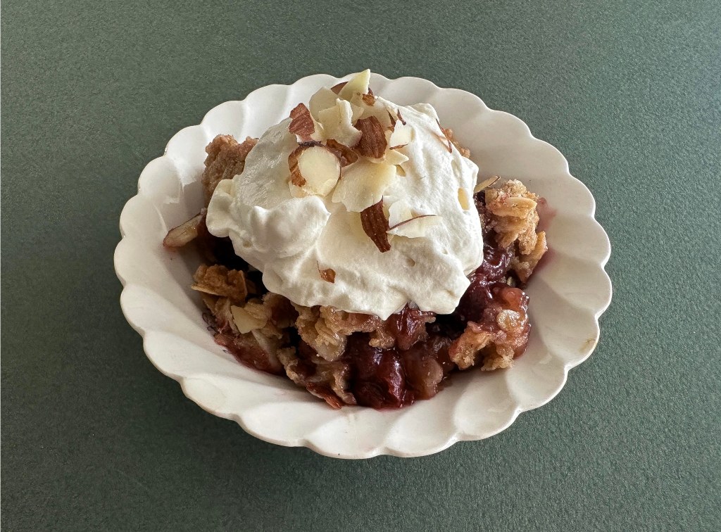 Mixed berry crisp topped with whipped cream and sliced almonds in a white scalloped bowl